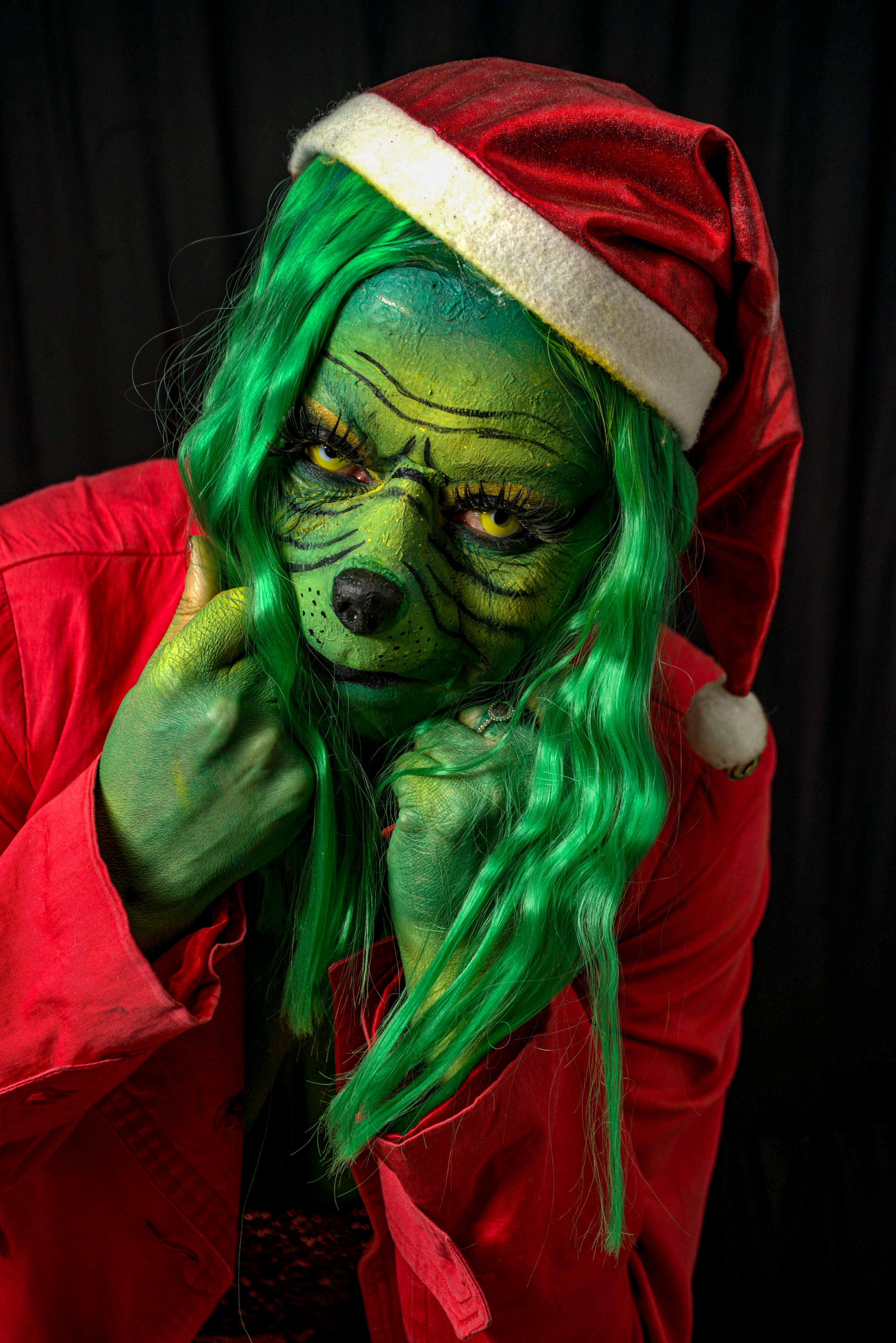 A person with green face paint wearing a red Santa hat and coat, embodying a holiday character in a playful pose.