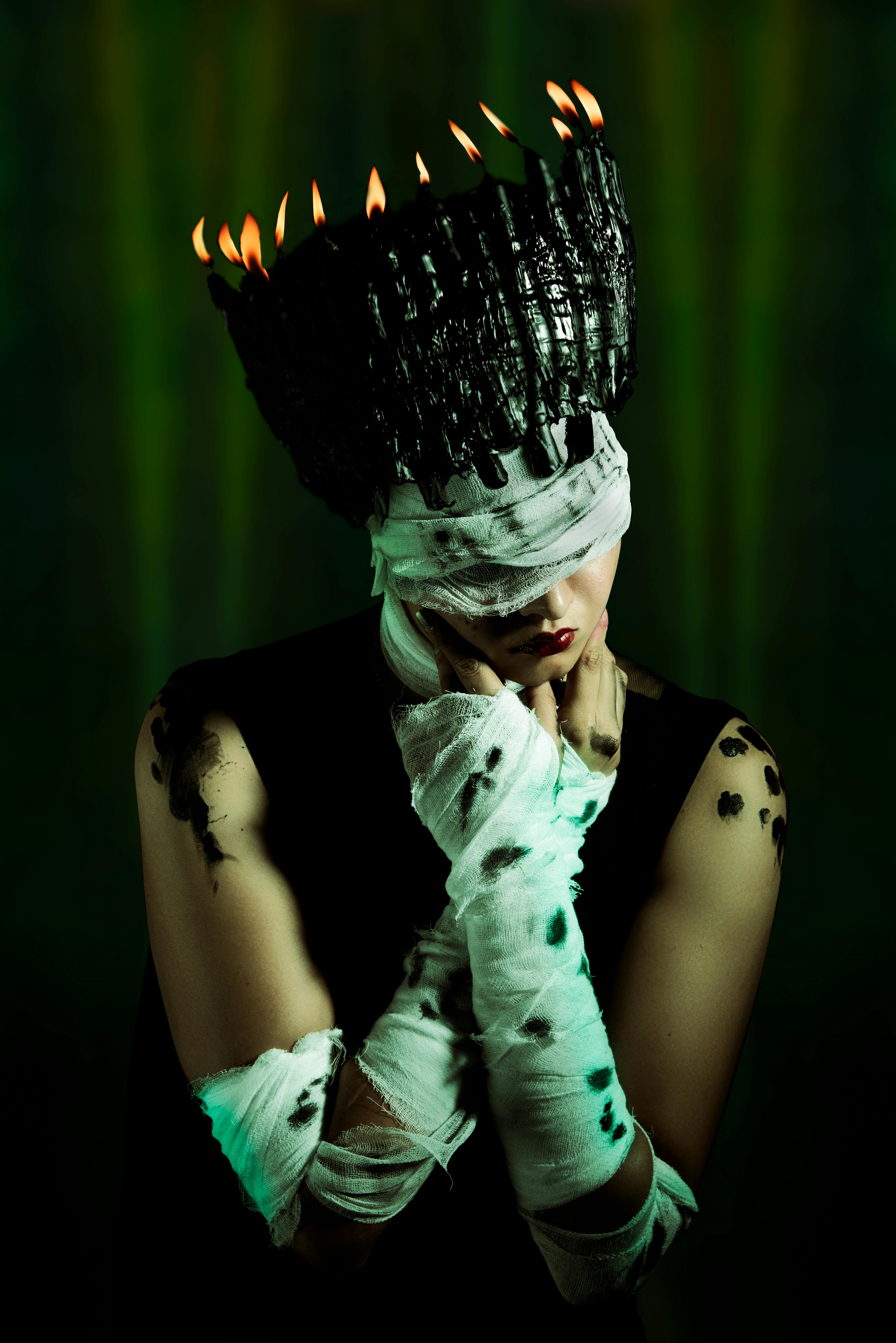 An enigmatic figure wearing a dark candle crown with bandaged eyes and arms, green backdrop.