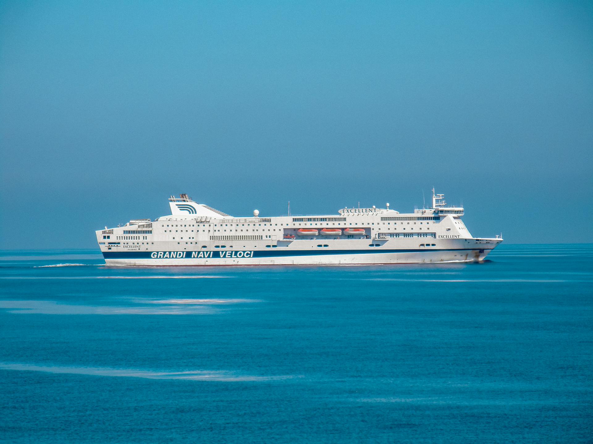 Large cruise ship 'Grandi Navi Veloci' sailing on serene blue waters under a clear sky.
