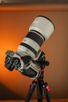 Close-up of a camera with a telephoto lens mounted on a tripod, against a warm background.
