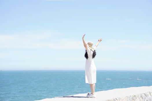 Woman in a white dress and hat raises arms joyfully by the ocean on a sunny day.