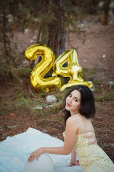 Young woman in floral dress celebrates 24th birthday with balloons in a natural setting.
