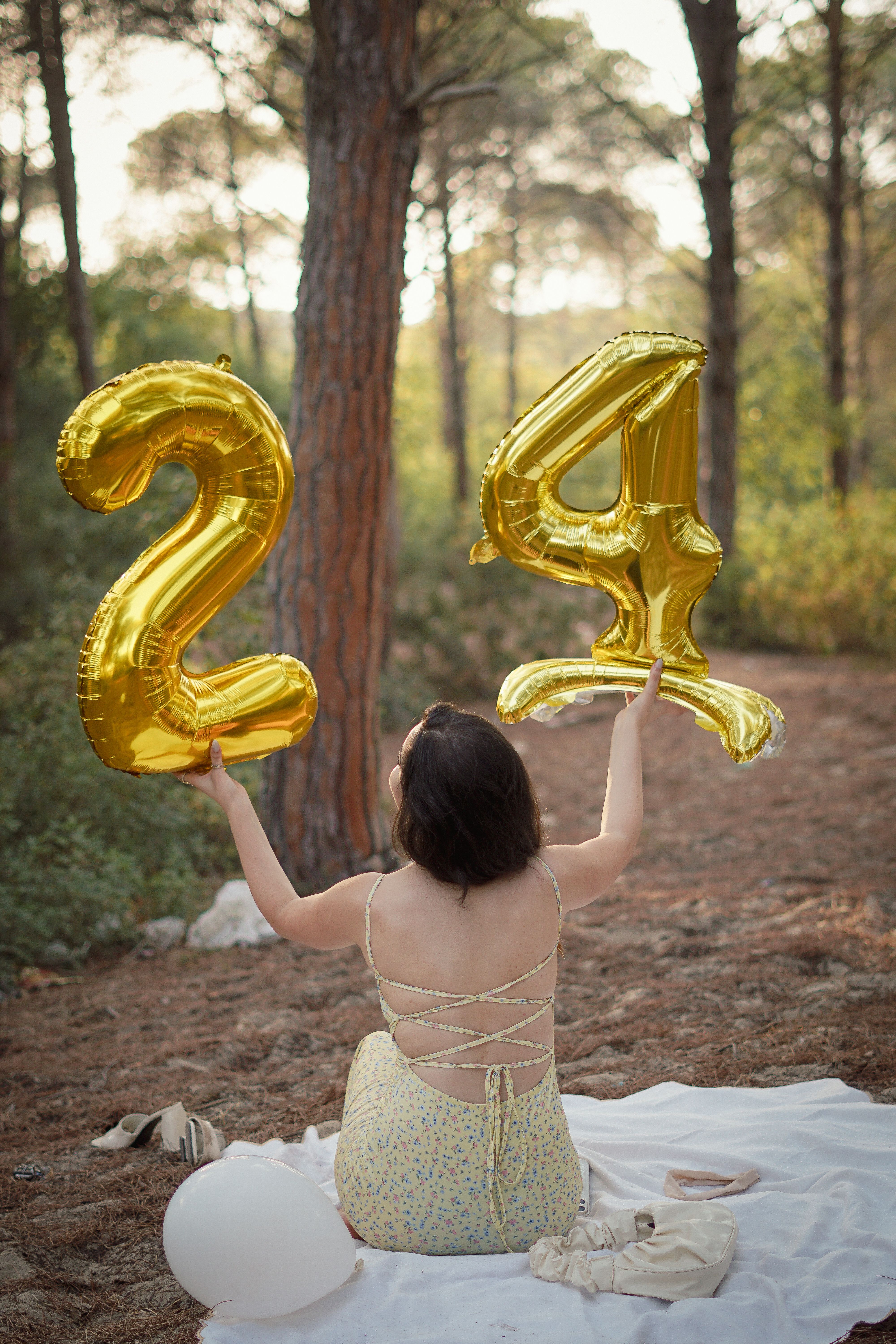 A woman in a summer dress celebrates holding large gold '24' balloons in a forest setting.