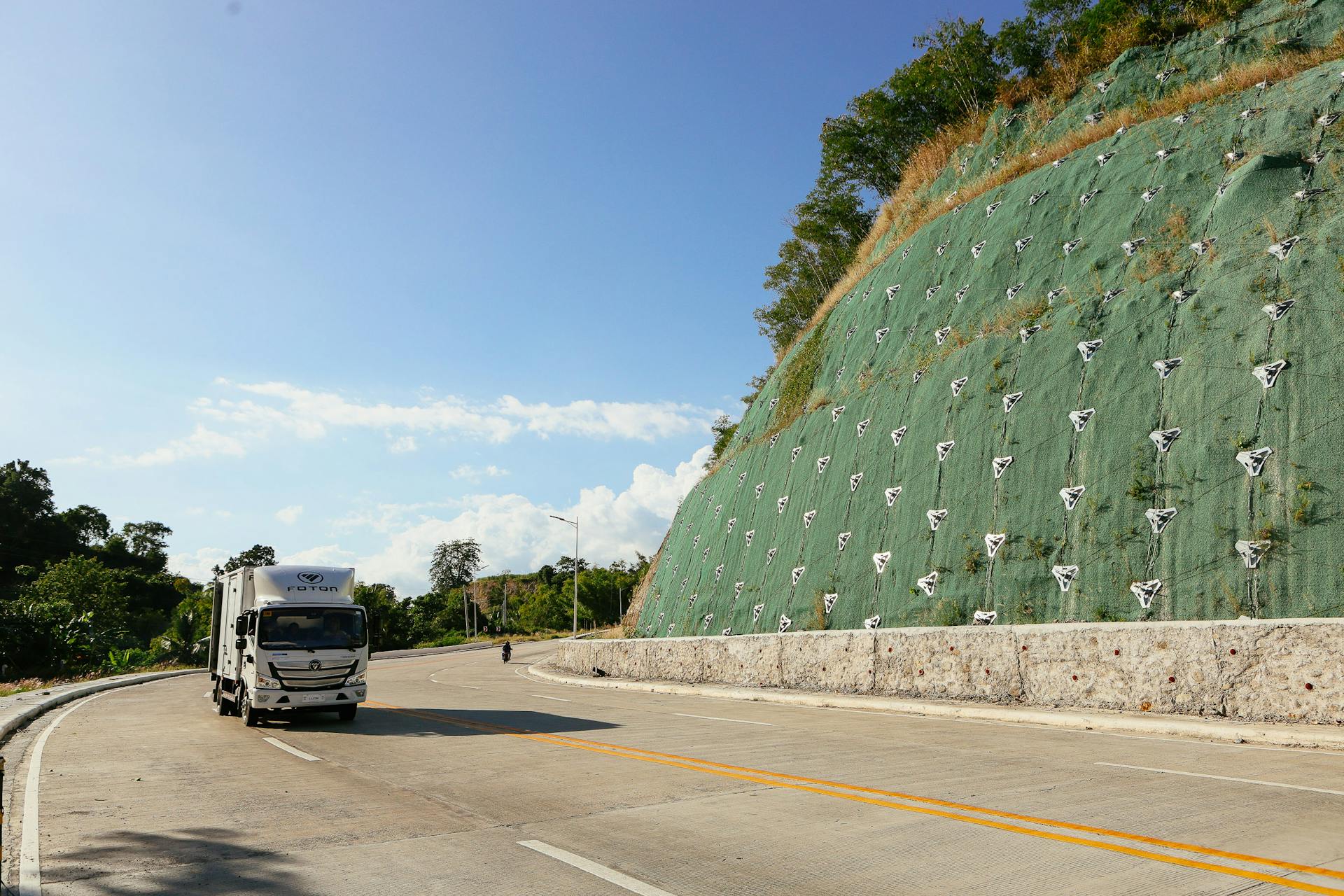 A delivery truck travels on a highway beside a hill with netting reinforcement.