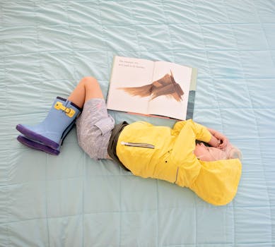A child in a yellow raincoat and boots relaxes with a book on a light blue quilted bedspread.