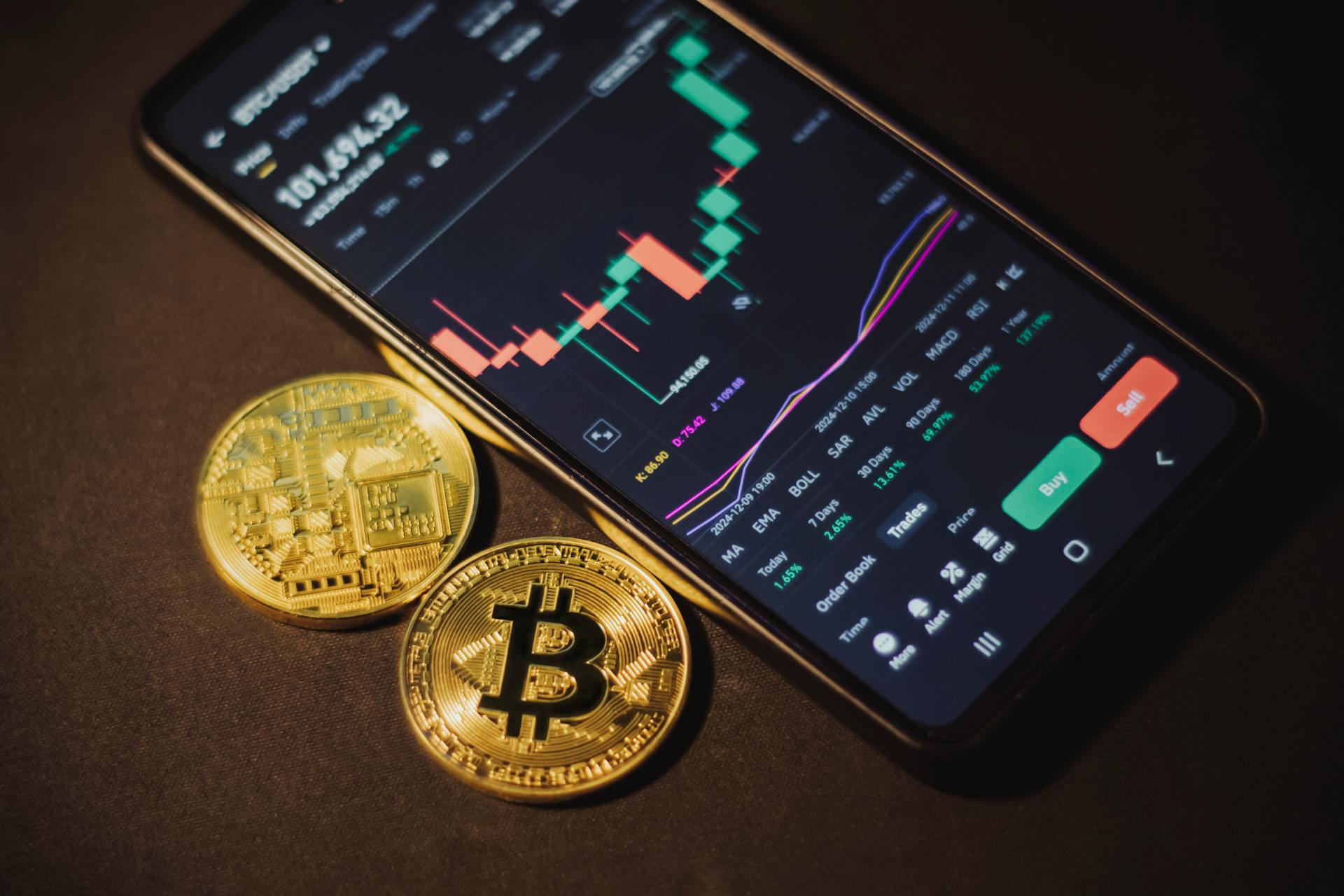 Smartphone displaying trading app with Bitcoin coins on a dark surface.