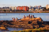 Group of people enjoying a sunny day by the river with a cityscape backdrop, capturing a vibrant urban lifestyle.