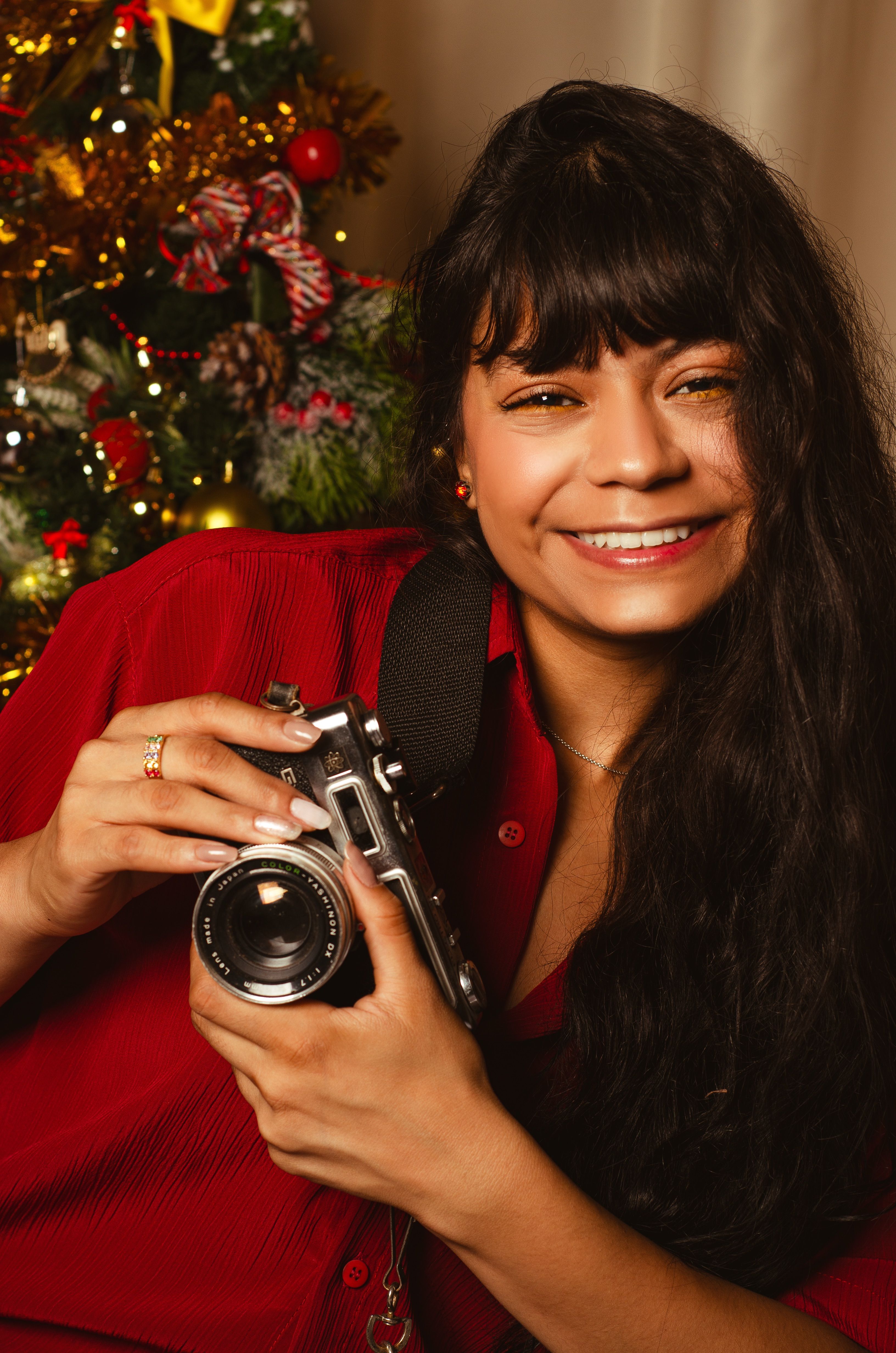 A joyful woman holding a camera, posing beside a decorated Christmas tree indoors.