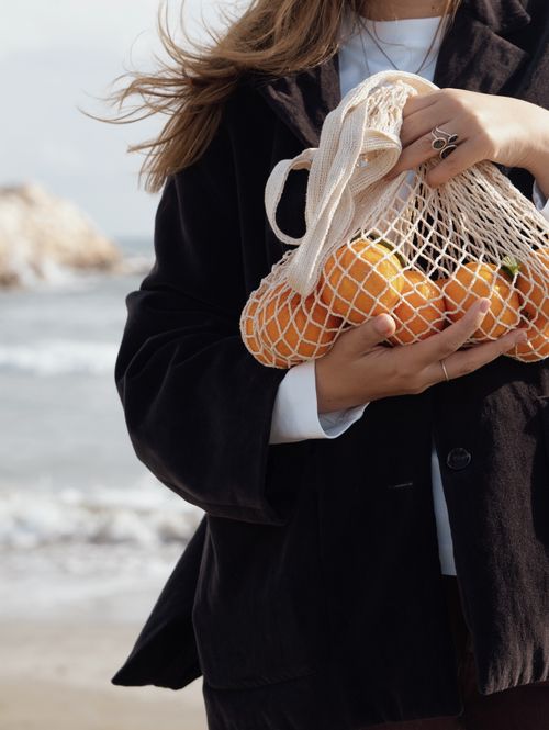 Free A woman carrying a mesh bag filled with oranges at the seaside beach, emphasizing sustainability and nature. Stock Photo