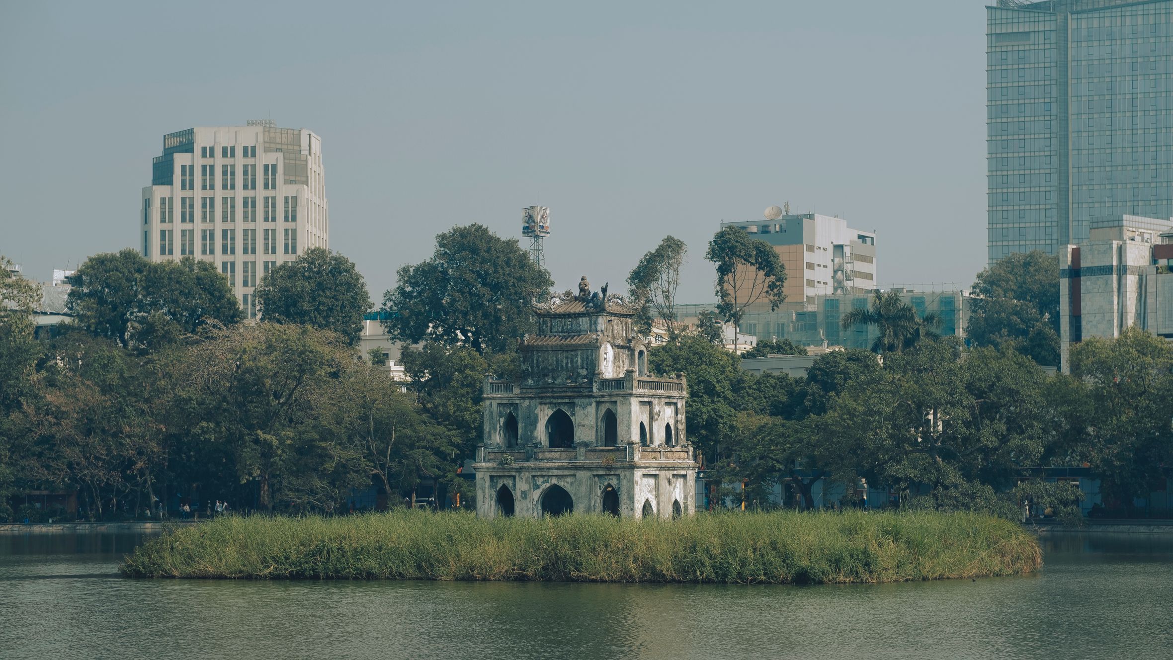 Scenic view of Turtle Tower in Hanoi's Hoan Kiem Lake surrounded by urban buildings.
