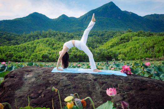 Woman practicing yoga on a rock in vibrant lotus field with scenic mountains in Hội An, Vietnam.