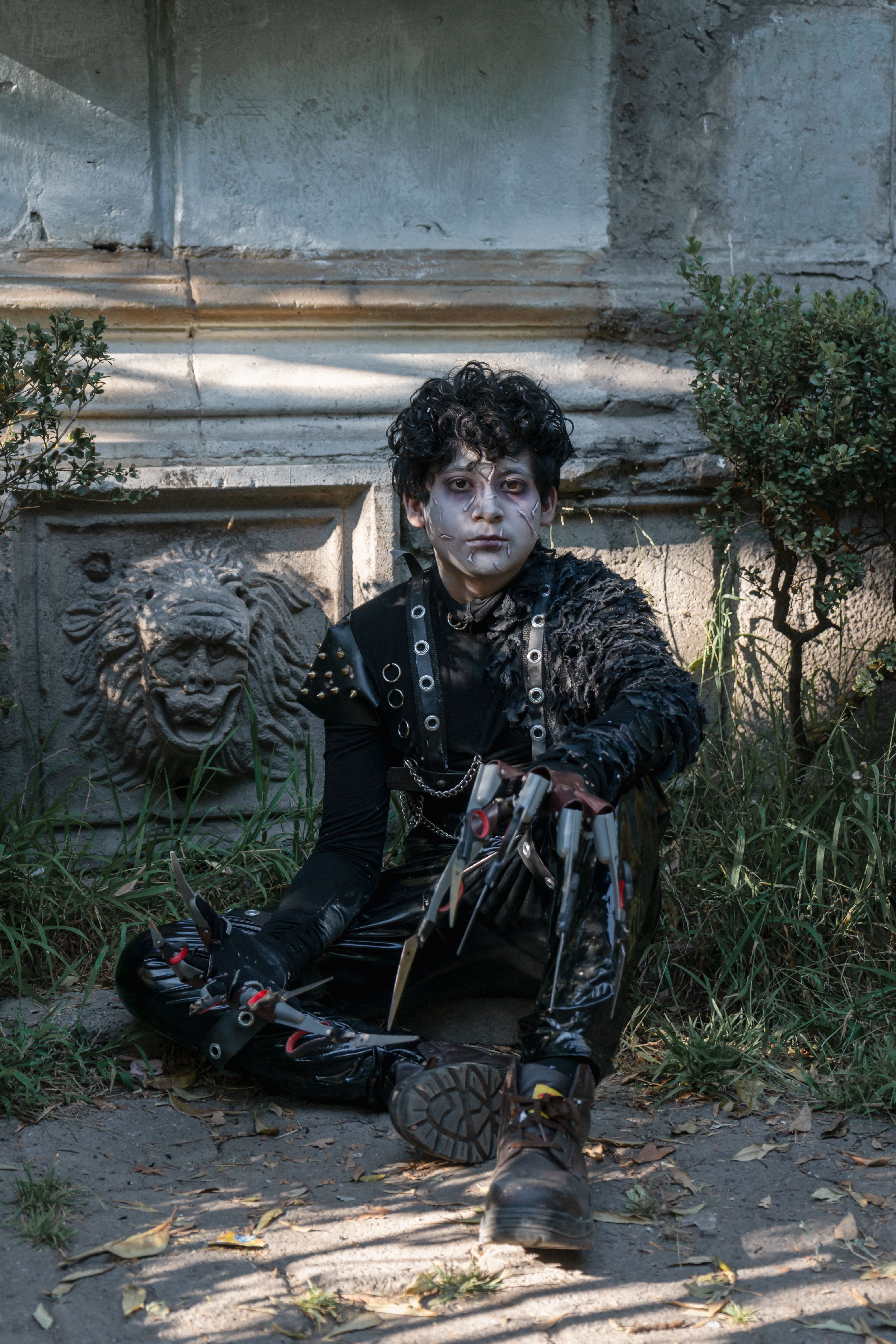 A person in gothic cosplay with unique makeup and attire sitting amidst ancient garden sculptures.