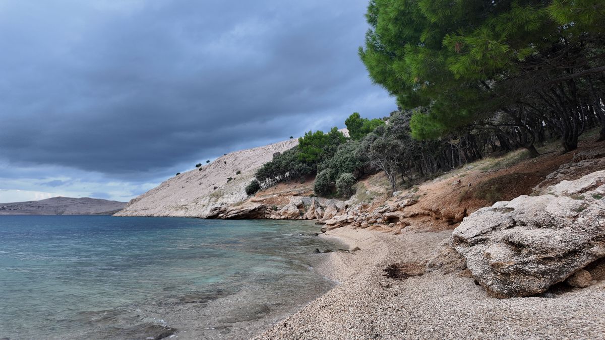 Plajă cu pietriș pe insula Pag, cu coasta împădurită coborând spre apa limpede
