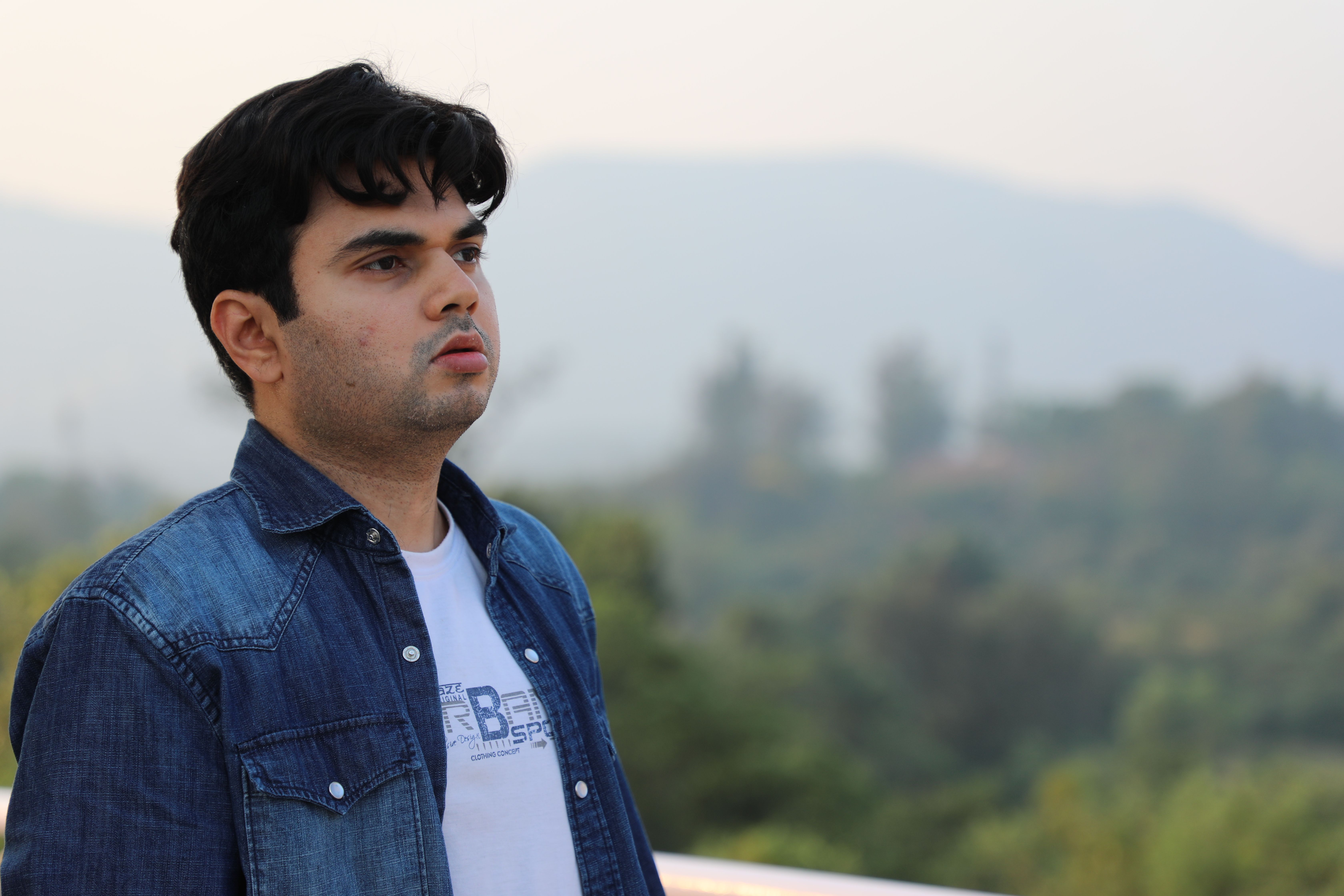 Young man wearing a denim jacket gazing outdoors with a blurred natural landscape behind.