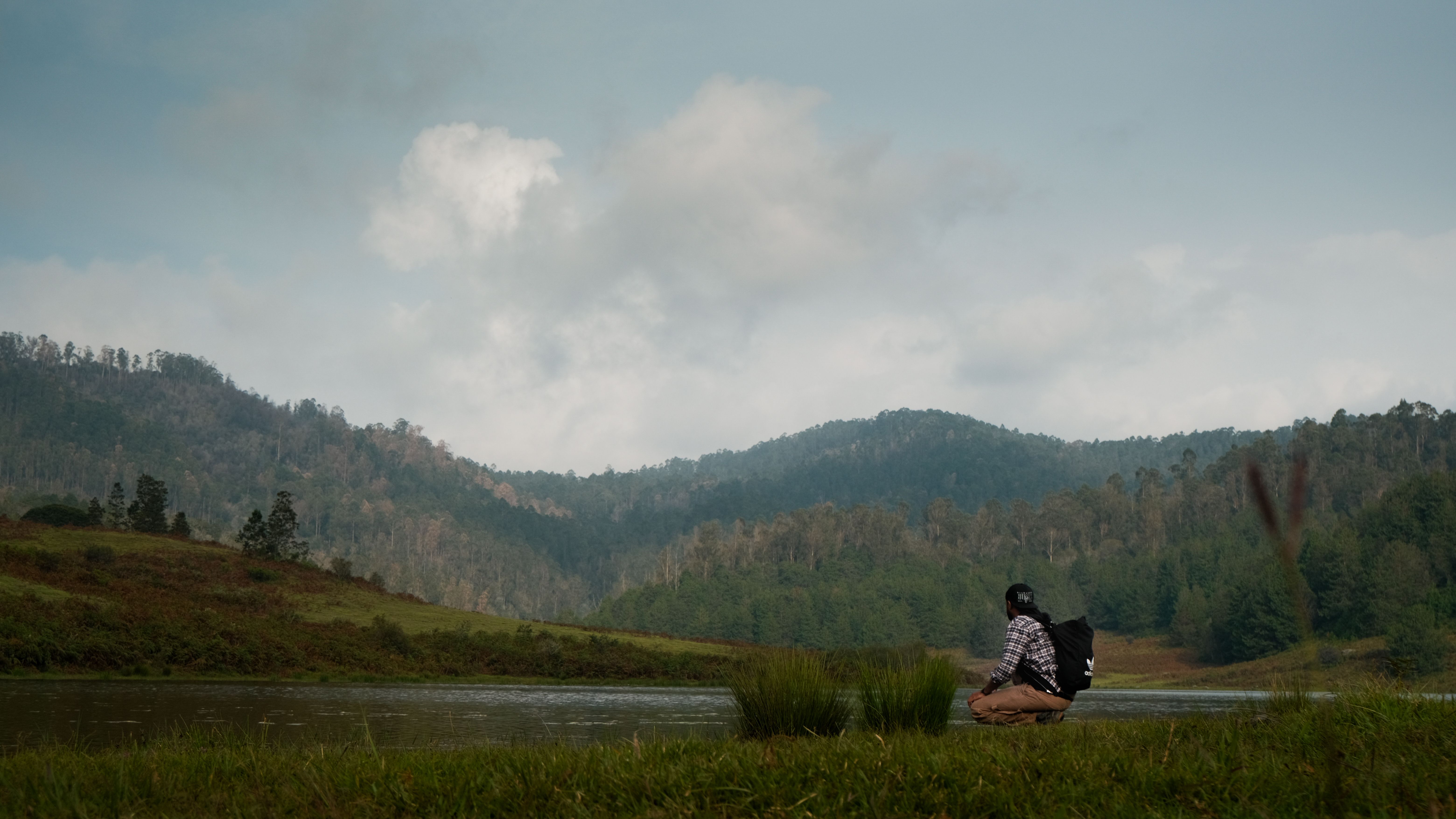 A lone hiker sits by a tranquil mountain lake surrounded by lush greenery under a cloudy sky.
