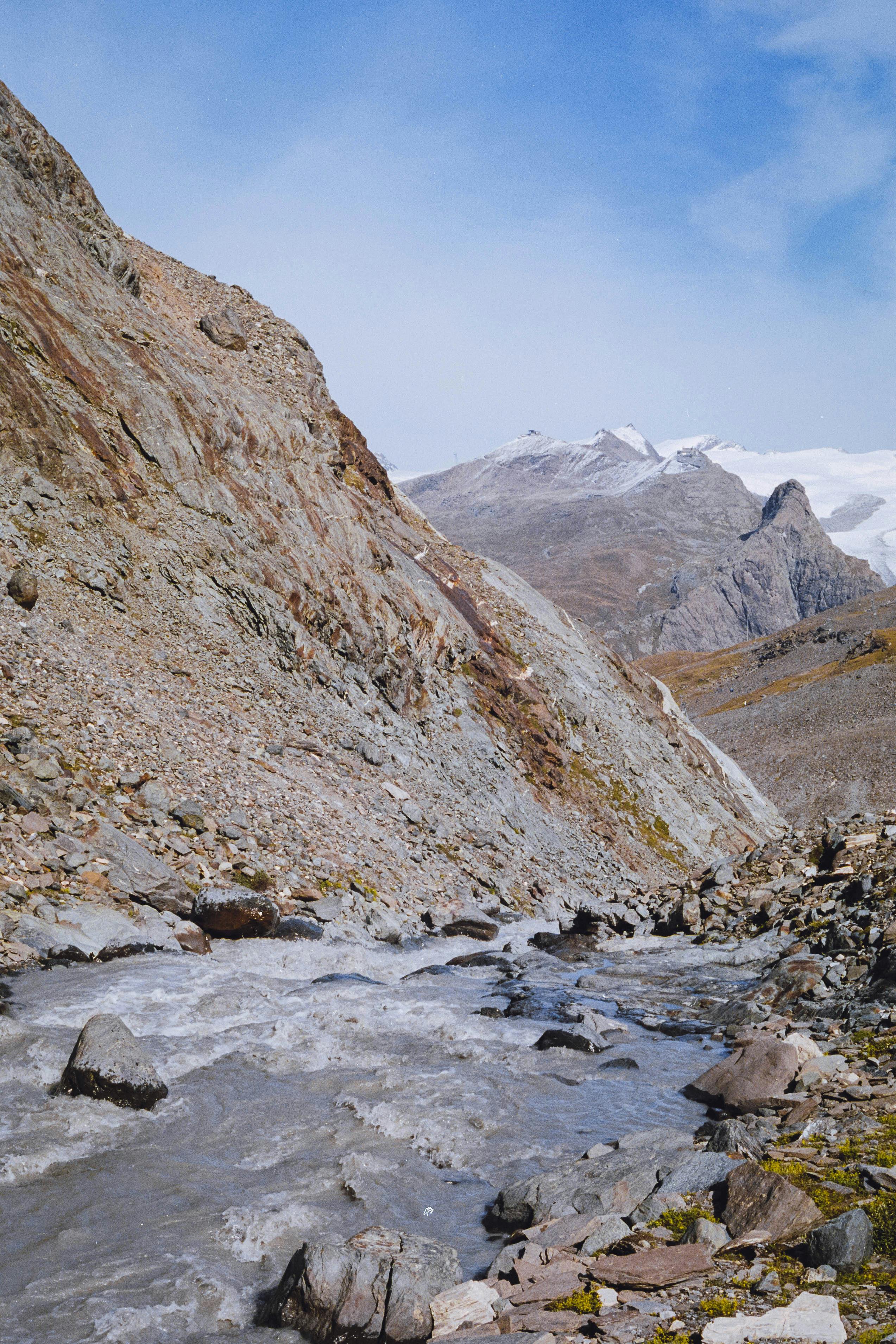 A peaceful mountain stream flows through a rocky valley with snow-capped peaks in the background.