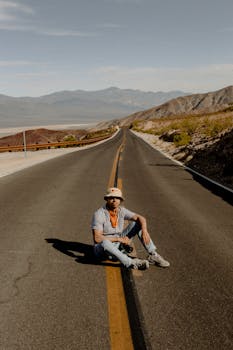 Casually dressed man sits on a deserted mountain highway on a sunny day.