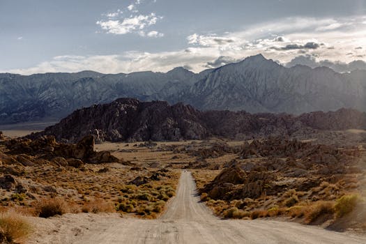 A scenic dirt road stretches towards a majestic mountain range under a cloudy sky.