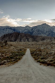 A tranquil dirt road leading towards majestic mountain ranges under a cloudy sky.