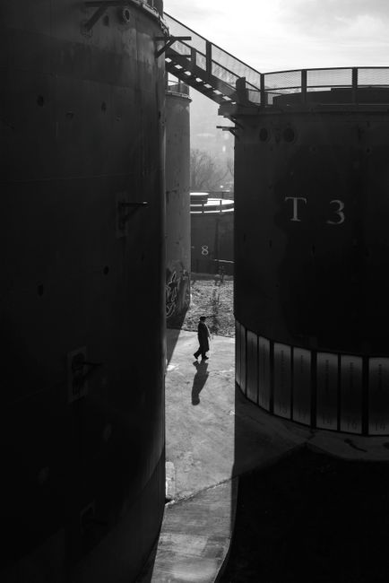 Black and white photo of a person walking between large industrial tanks, creating a striking silhouette.