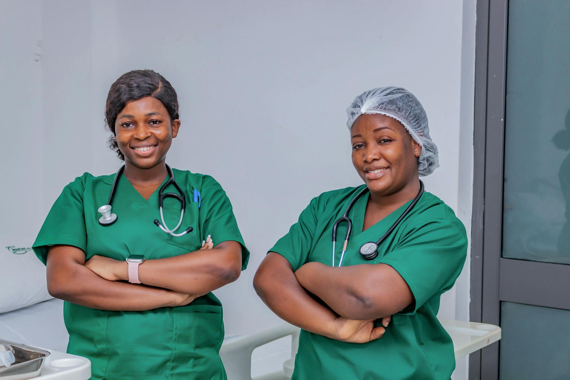 Two female nurses in green scrubs smiling confidently in a hospital room. Professional healthcare workers.