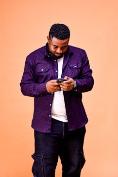 A man in a casual shirt focuses on his smartphone against an orange backdrop.