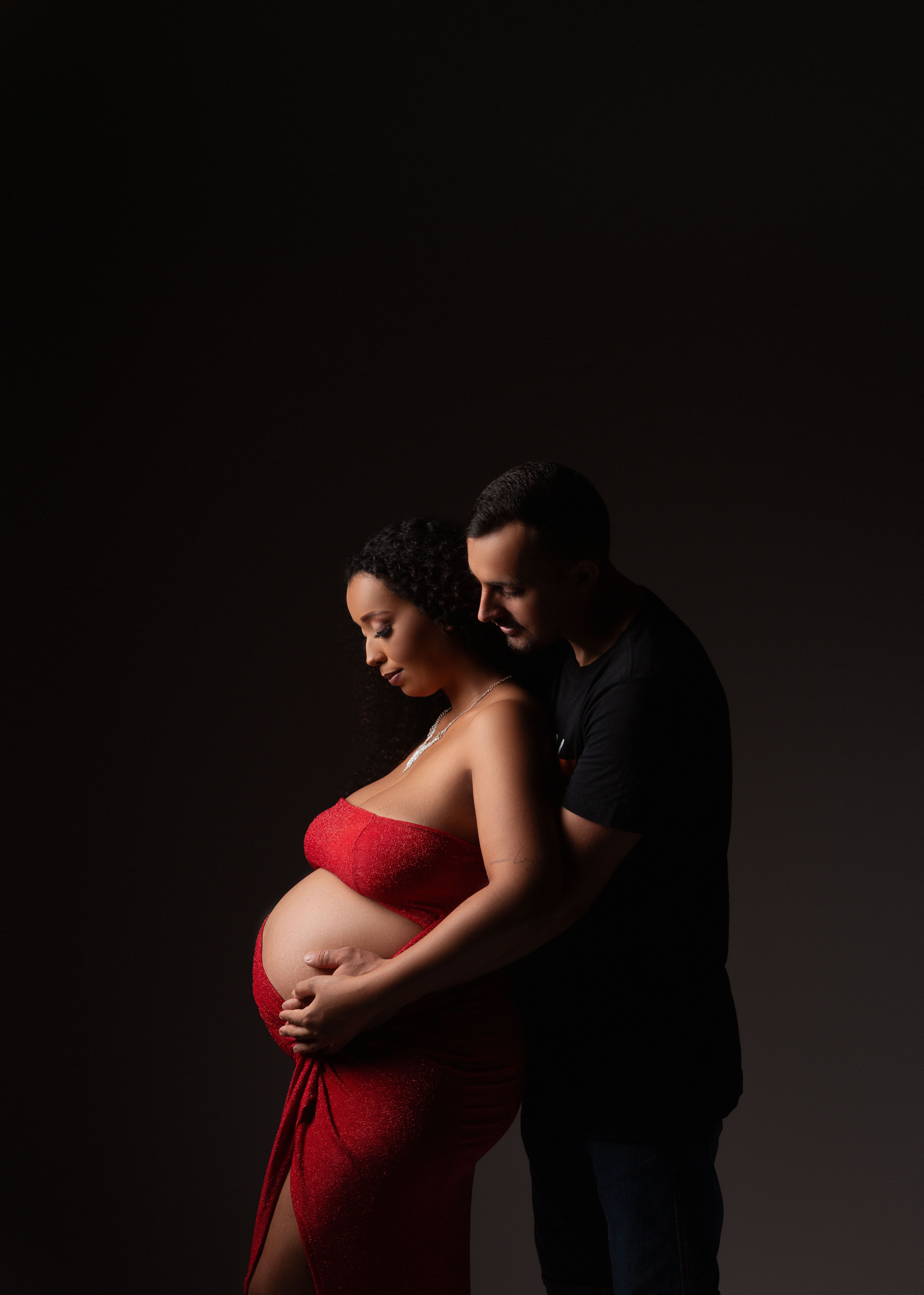 Pregnant woman in red dress with partner, intimate studio pose.