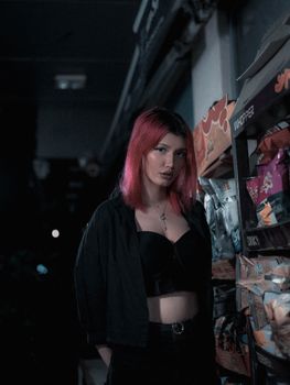 Stylish woman with pink hair poses in a dimly lit store aisle, showcasing urban fashion with cool-toned aesthetics.