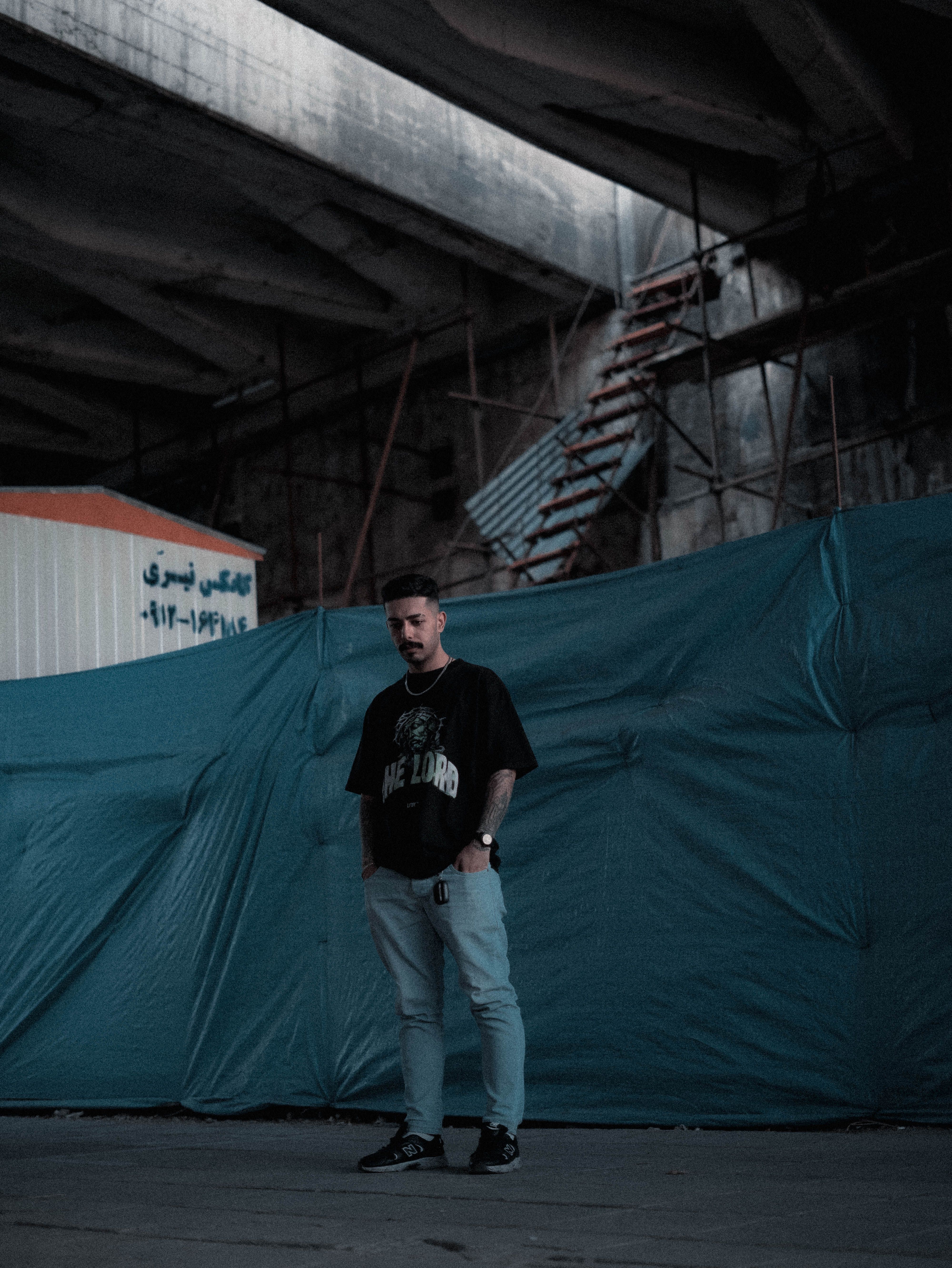 A man in casual attire stands in an industrial area with blue tarps and scaffolding, creating a moody atmosphere.