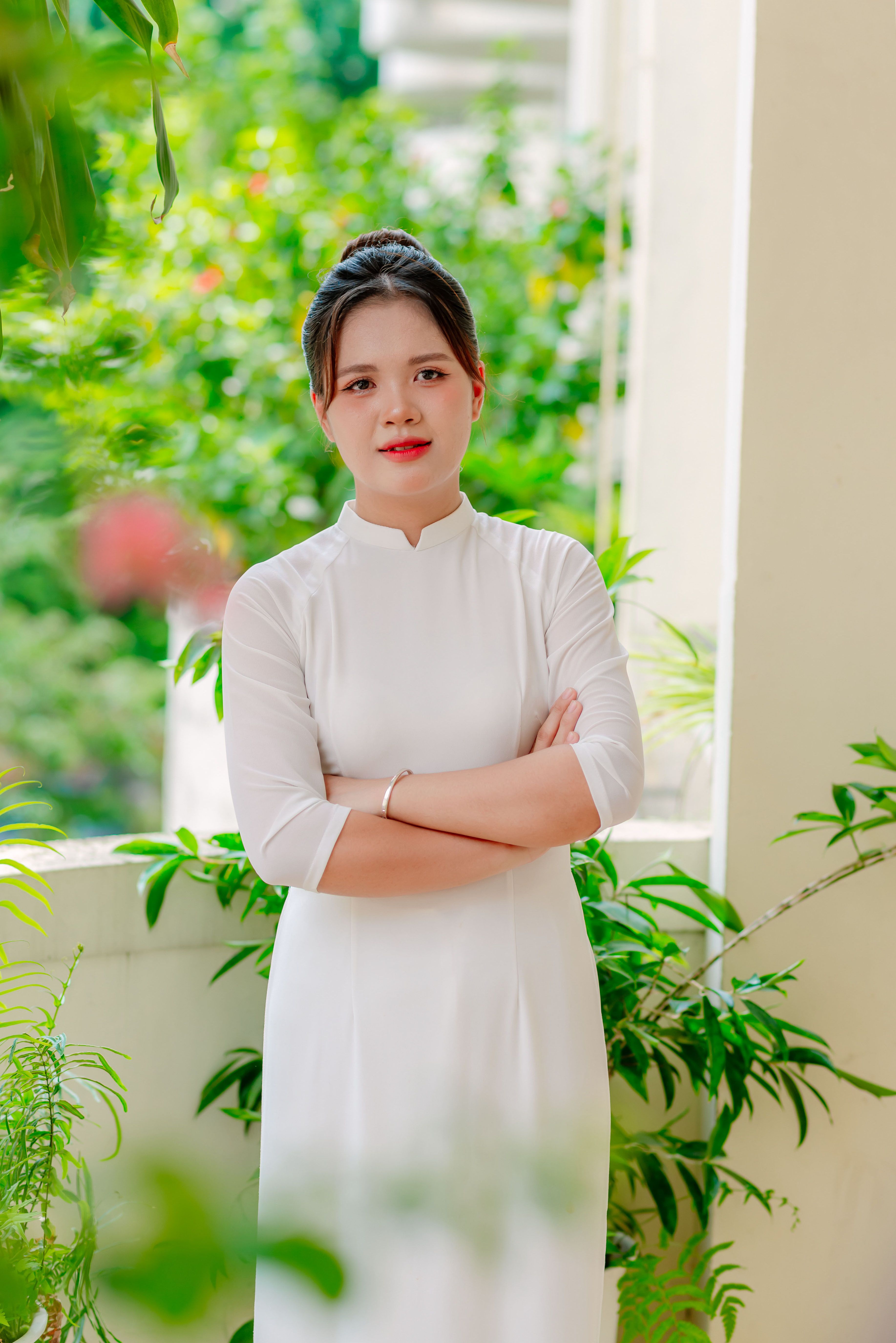 A woman standing outdoors in a white traditional dress with lush greenery in the background.