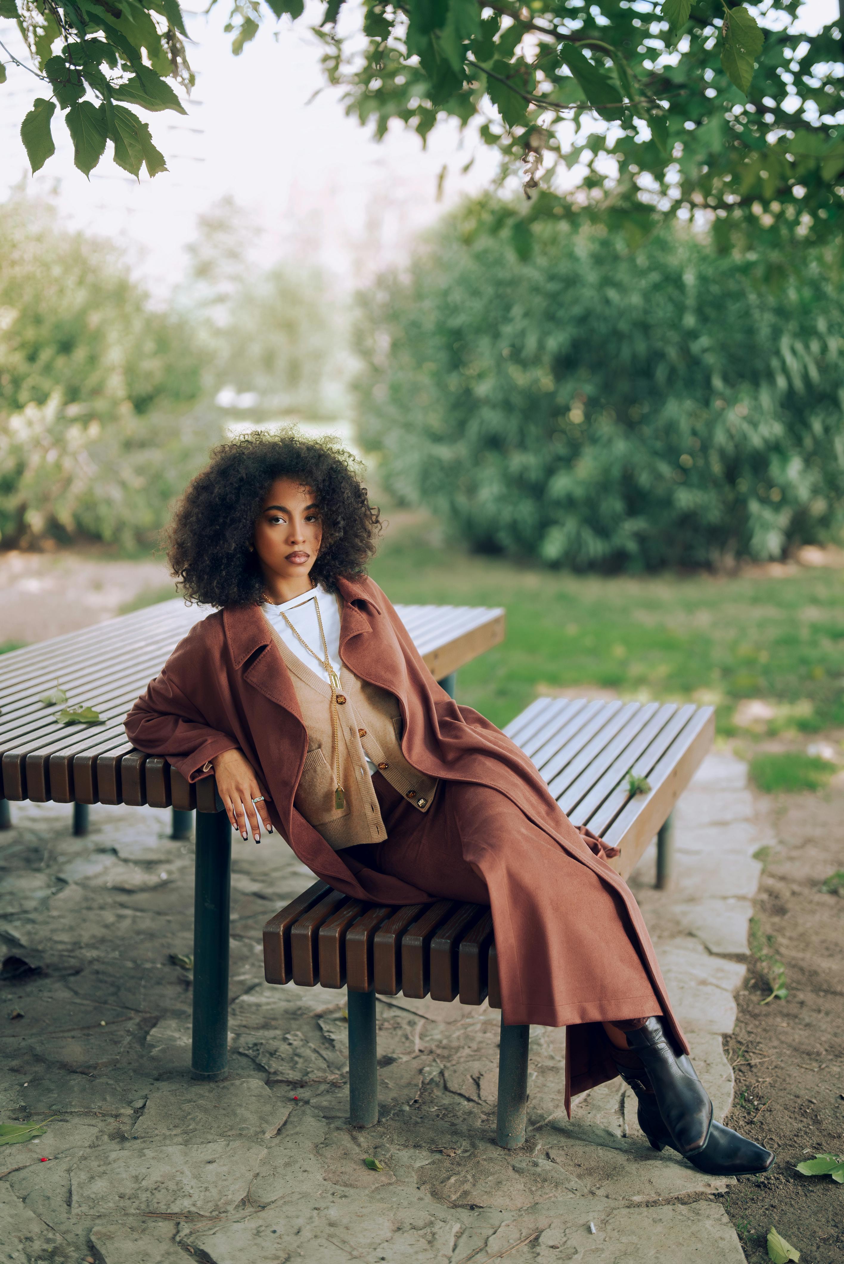 Stylish woman in brown coat sitting on a bench in a park setting.