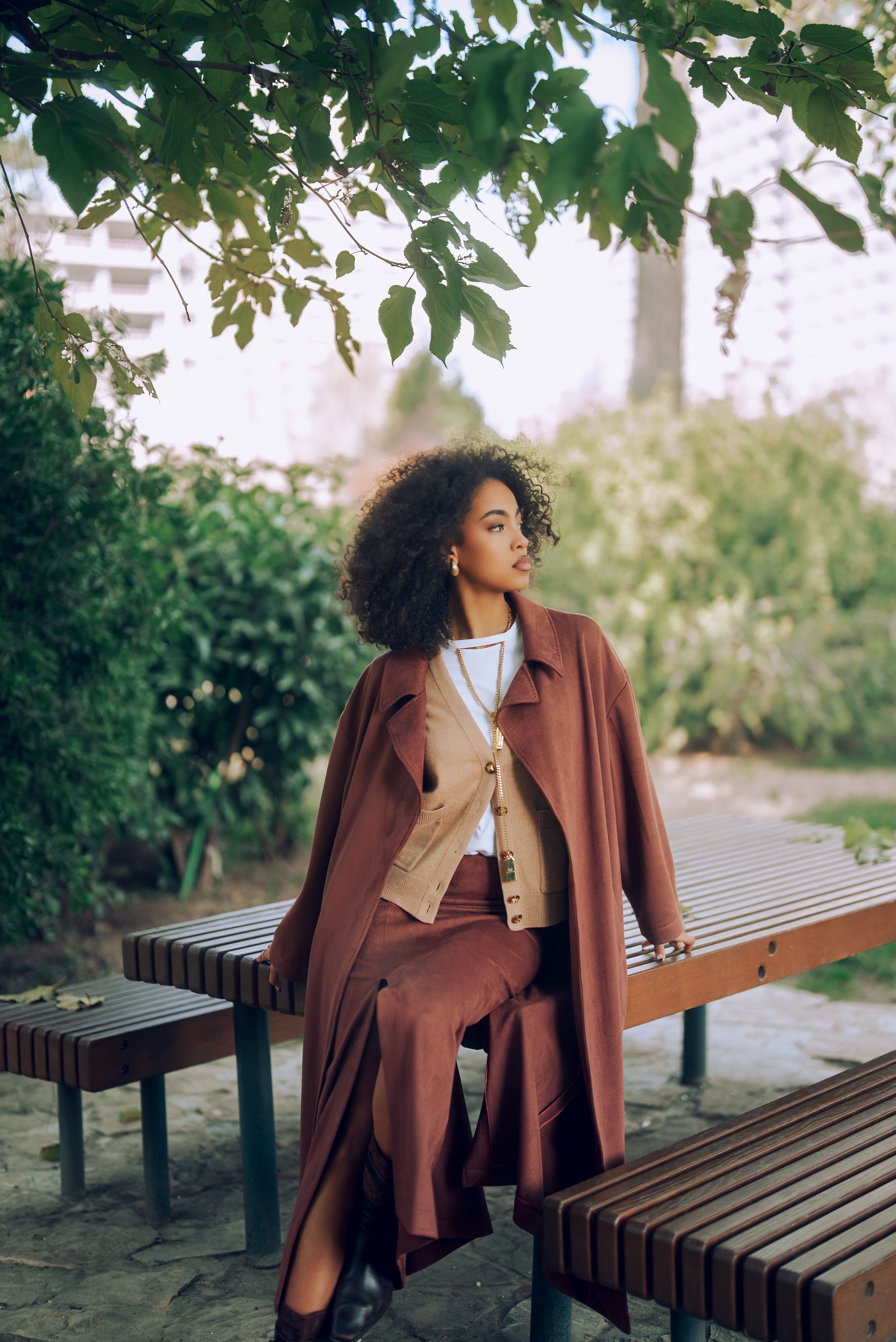 Stylish woman wearing autumn attire sitting on a park bench surrounded by green foliage.