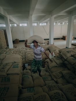 A man carrying a large sack amidst a warehouse full of burlap sacks, showcasing labor and storage.