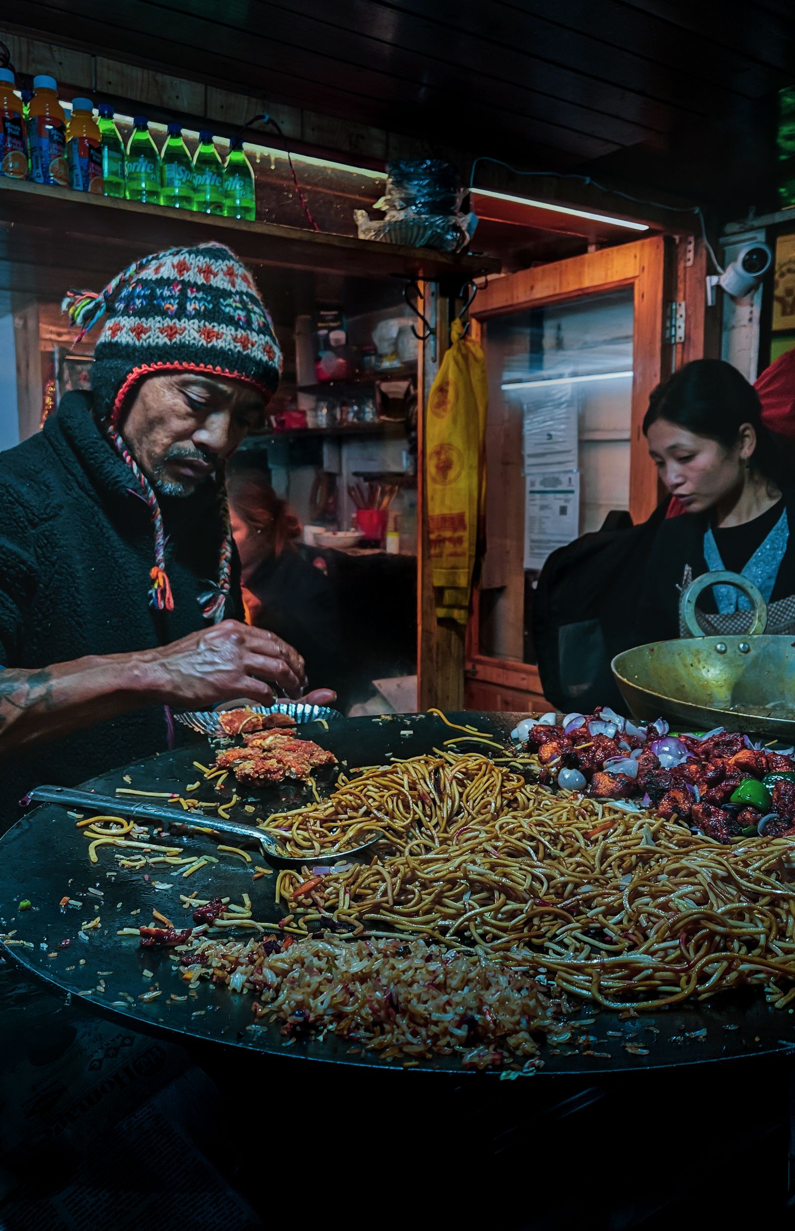 A cook in a woolly cap serving noodles and chilli fry from a wide griddle at a night market stall in Sikkim