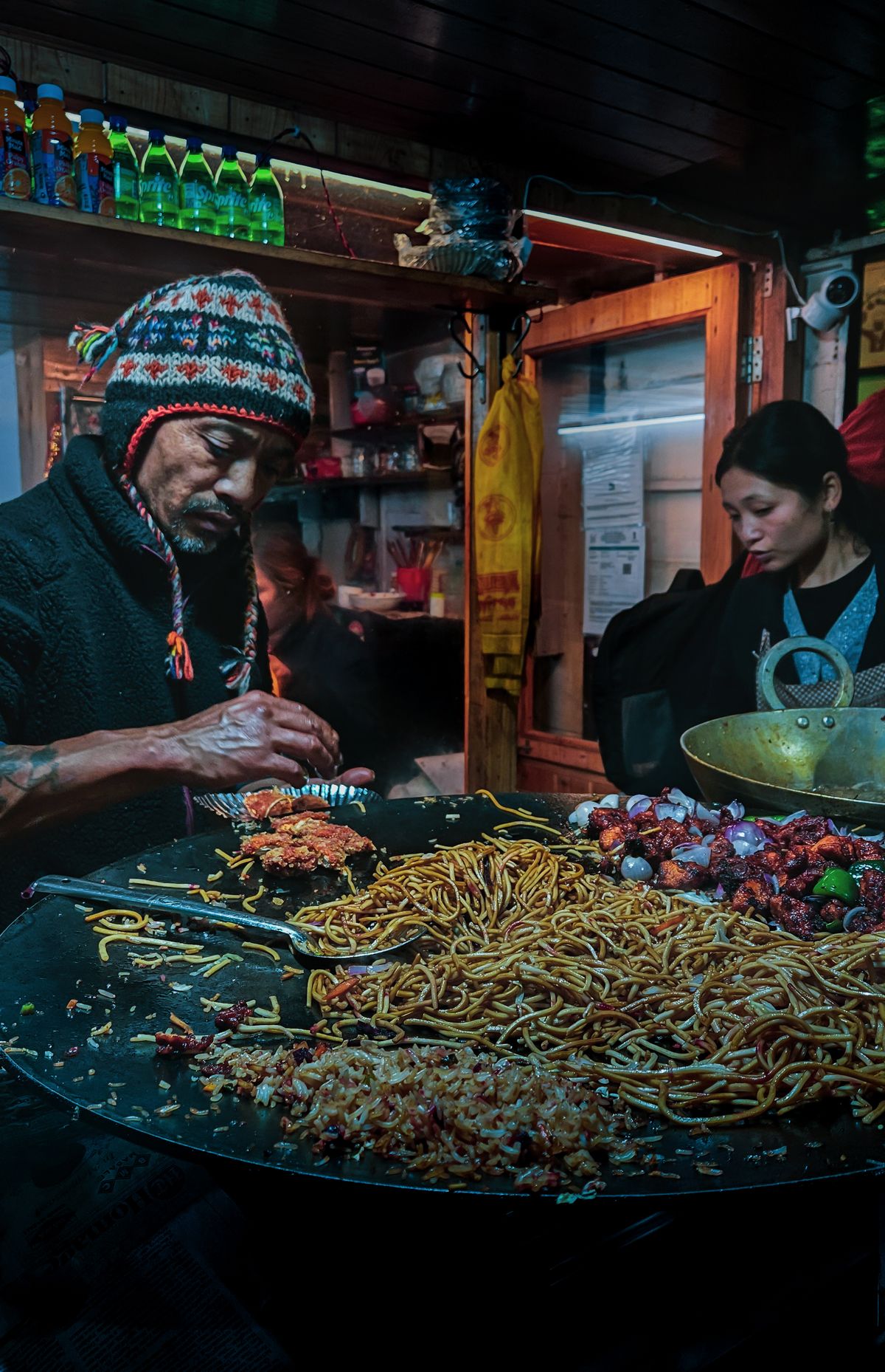 A cook in a woolly cap serving noodles and chilli fry from a wide griddle at a night market stall in Sikkim