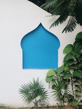 A minimalist blue arched window on a white wall surrounded by tropical leaves in Tulum, Mexico.