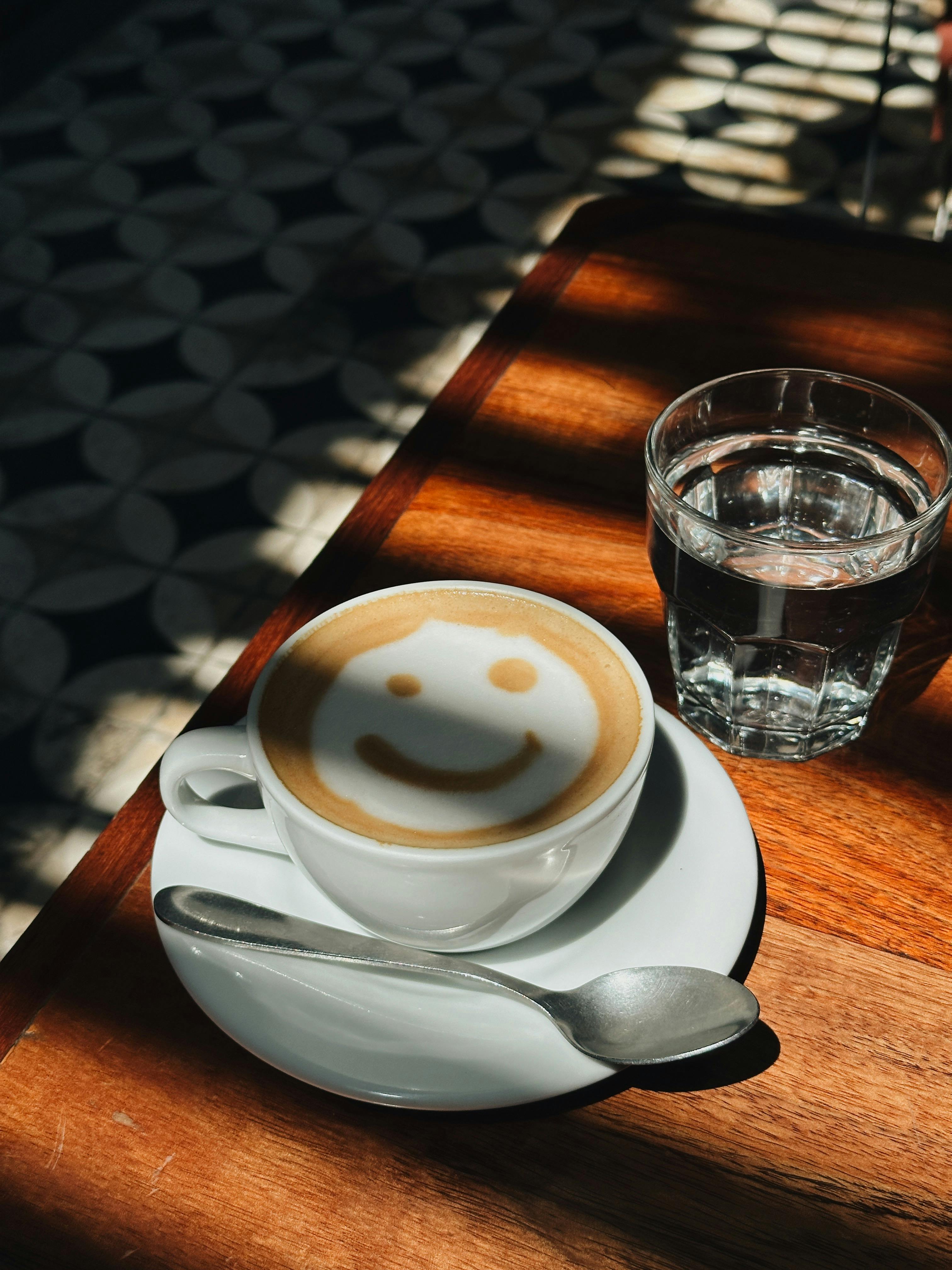 A cappuccino with smiley face latte art on a wooden table in natural light.
