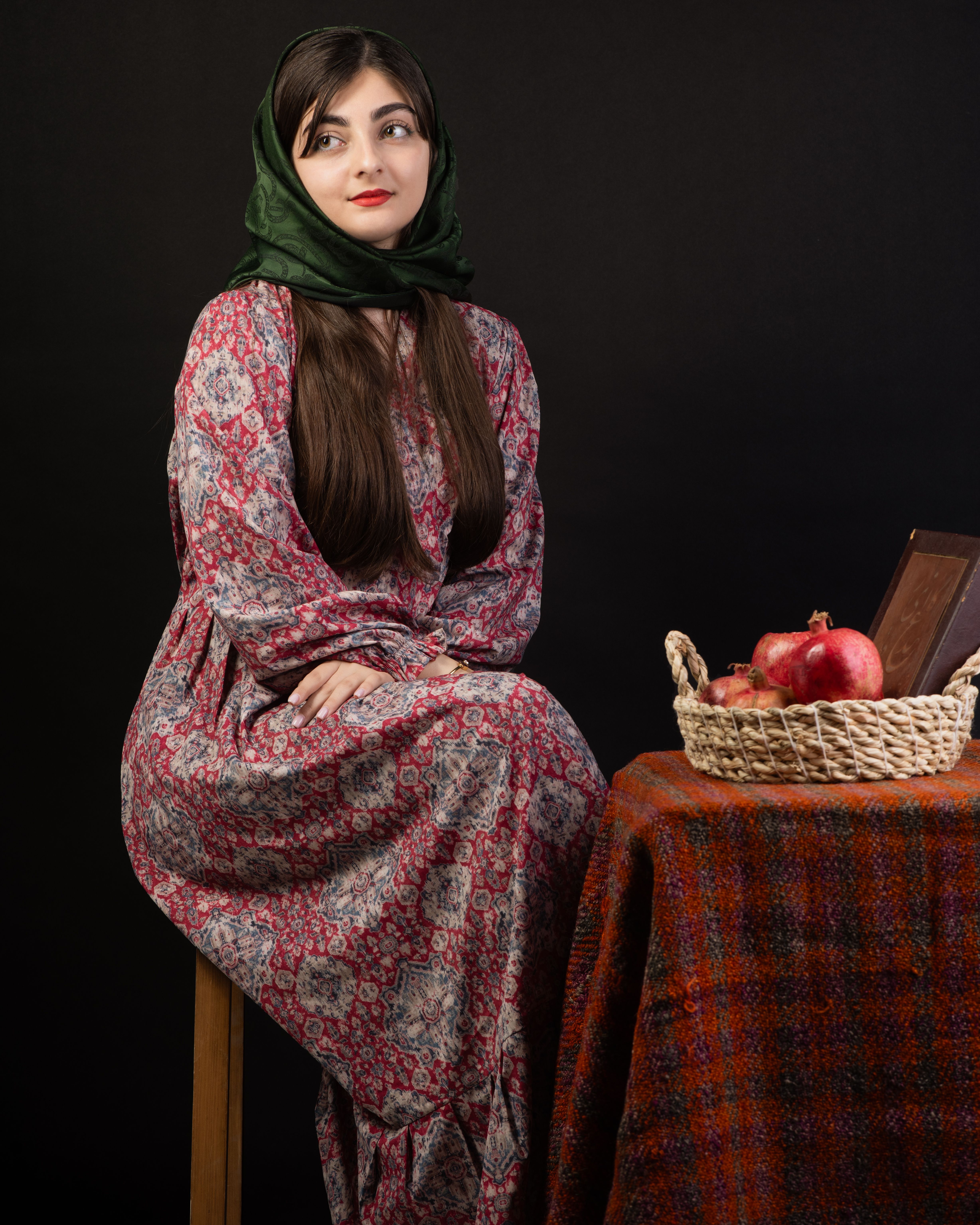 Elegant portrait of a woman in traditional dress with pomegranate basket on a patterned tablecloth.
