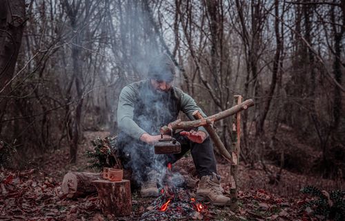 Free A man prepares food over a campfire in a dense forest, highlighting the essence of wilderness survival. Stock Photo