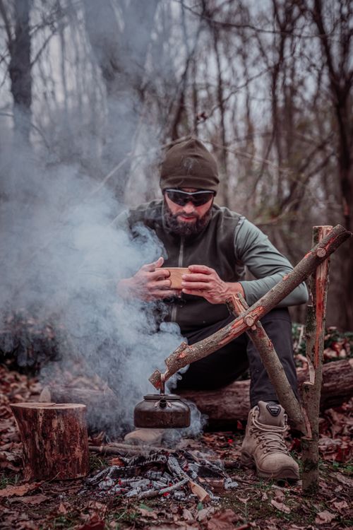 Free Bearded man drinking by a campfire in the woods, highlighting outdoor survival skills. Stock Photo