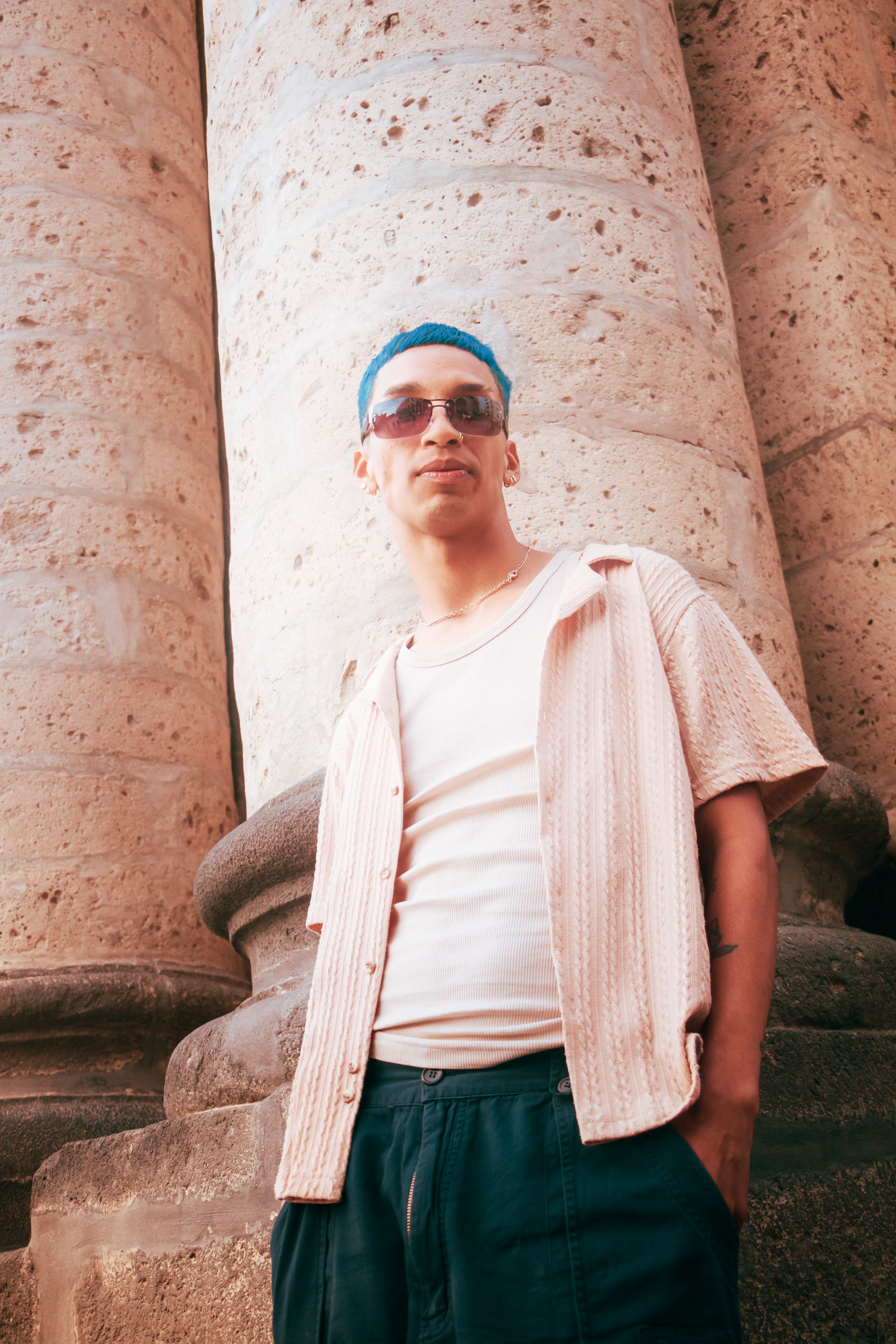 Young adult with blue hair poses confidently beside ancient stone columns.