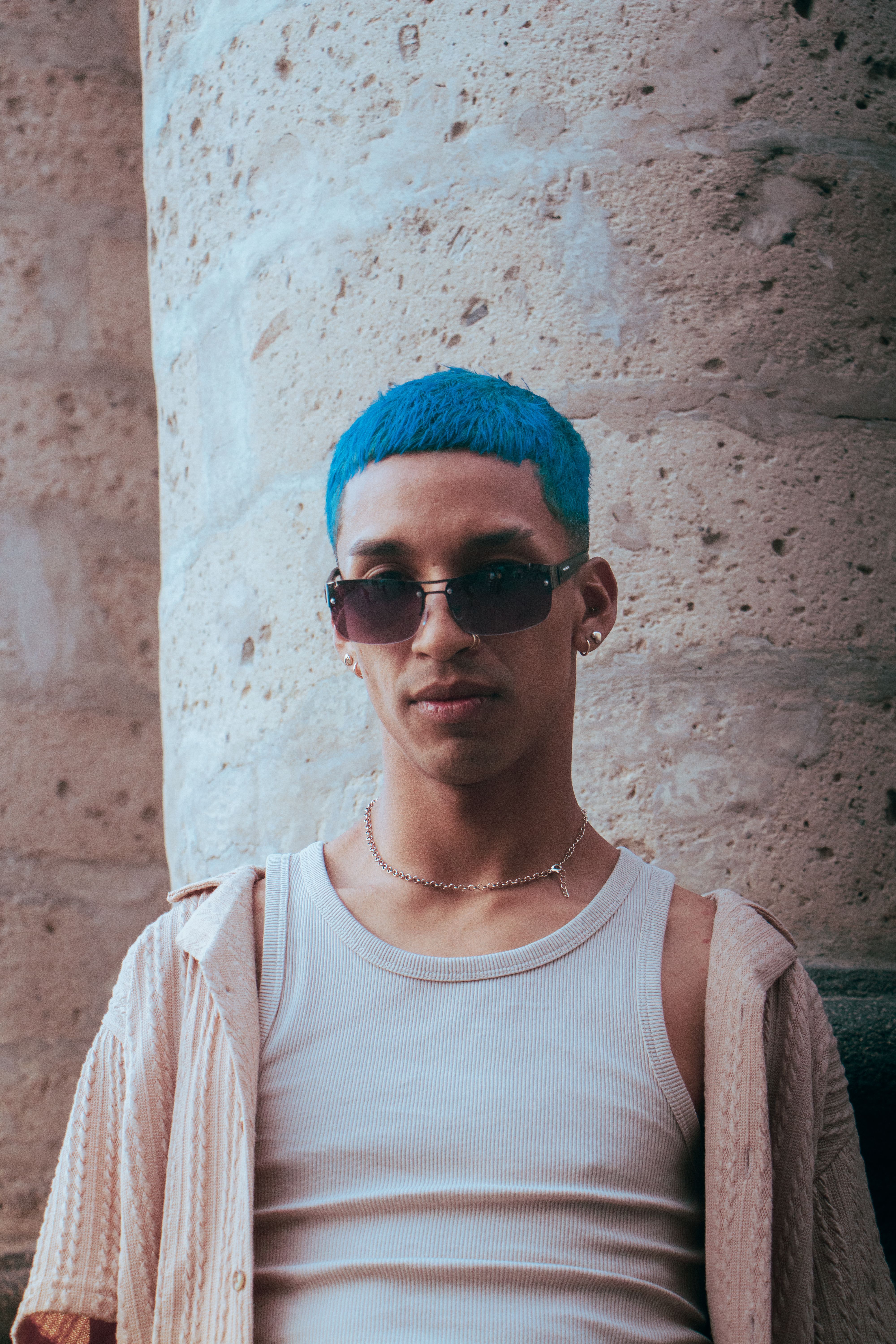 Stylish young man with blue hair and sunglasses standing against a stone wall outdoors.