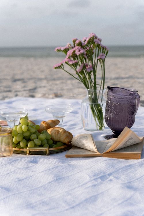 Free Beach picnic setup with grapes, croissants, lavender flowers, and a book. Stock Photo