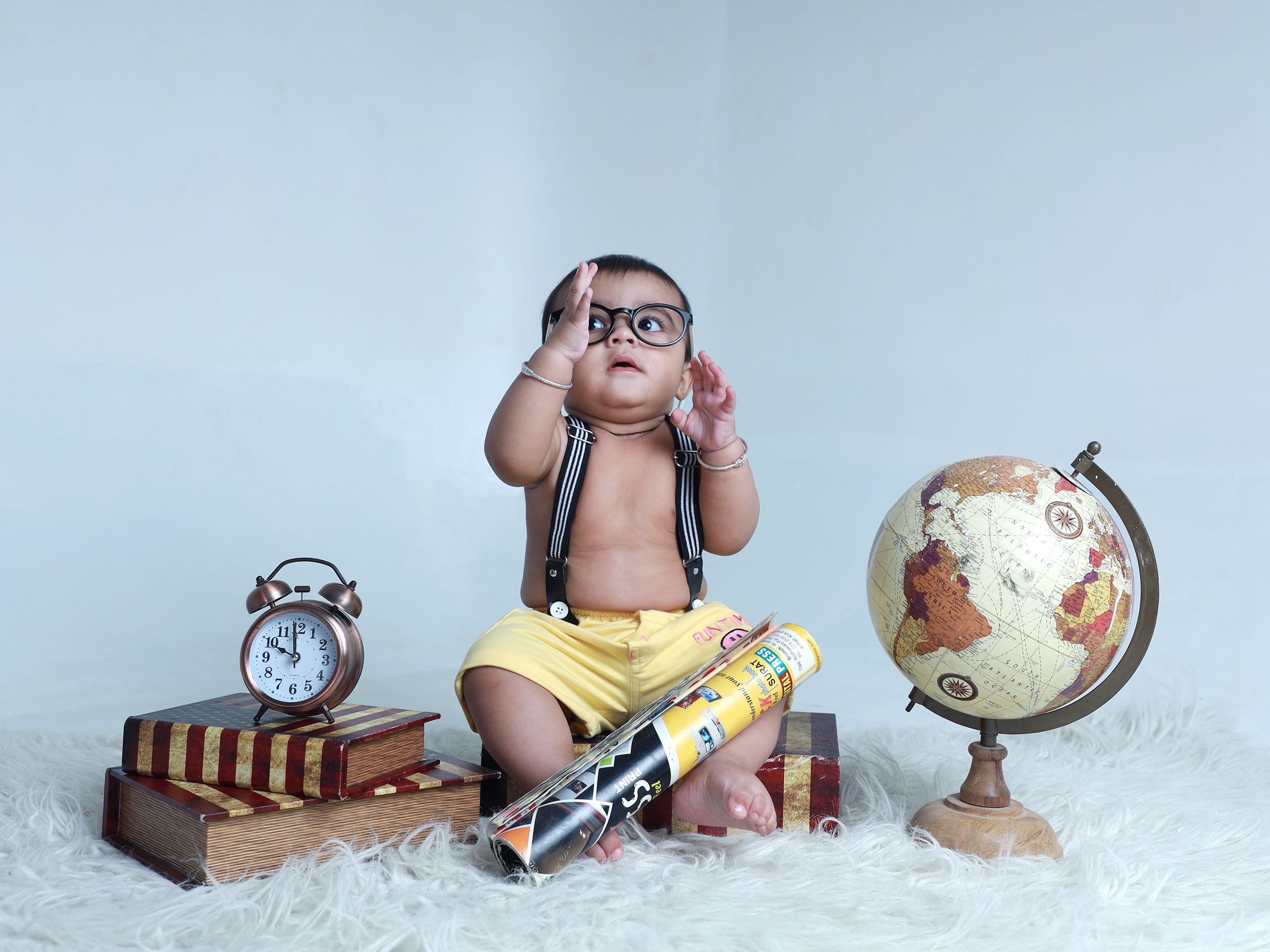 Charming baby in glasses on a cozy rug with books, globe, and vintage clock.