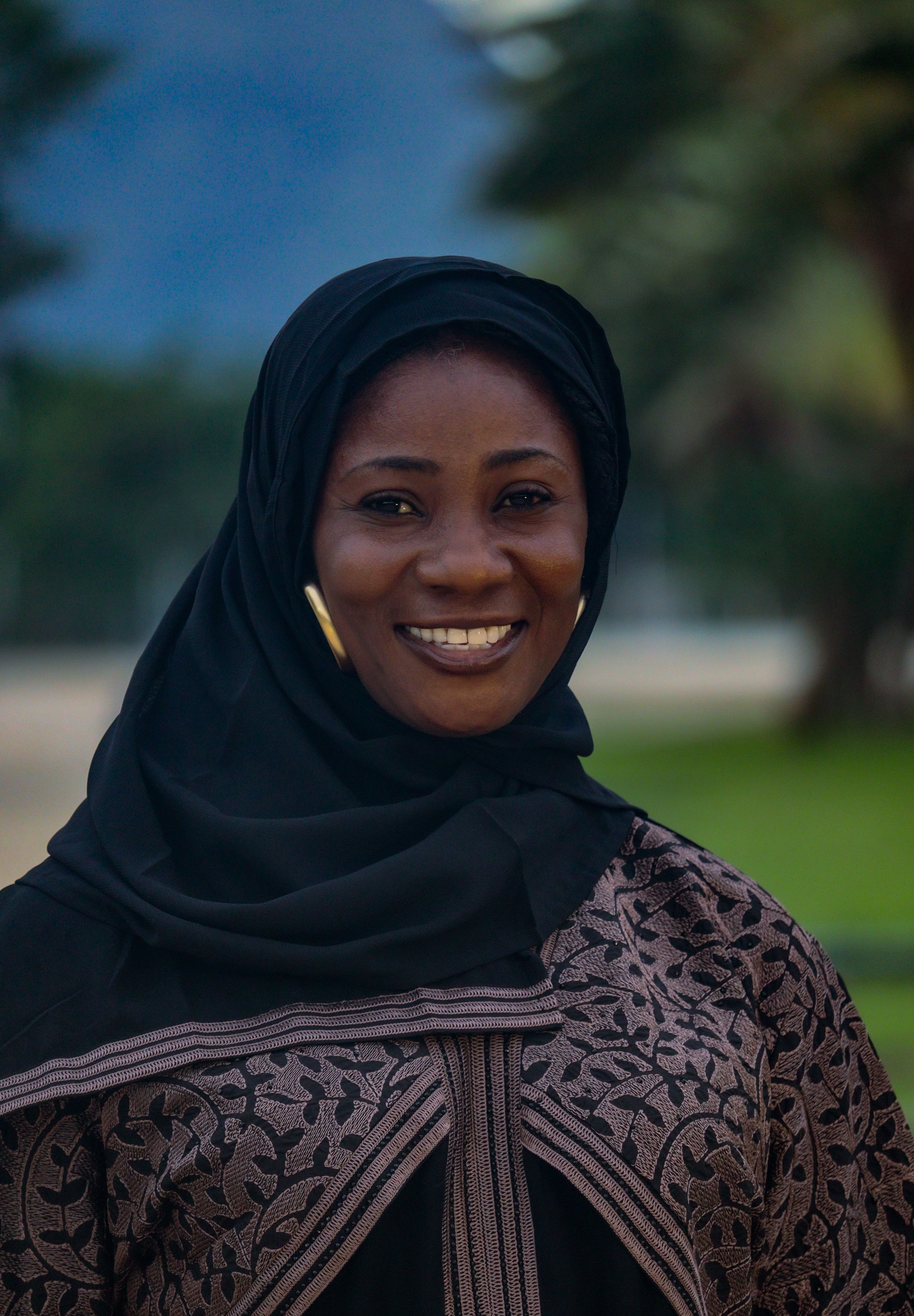 A cheerful woman wearing a traditional hijab and abaya, smiling warmly in an outdoor setting.