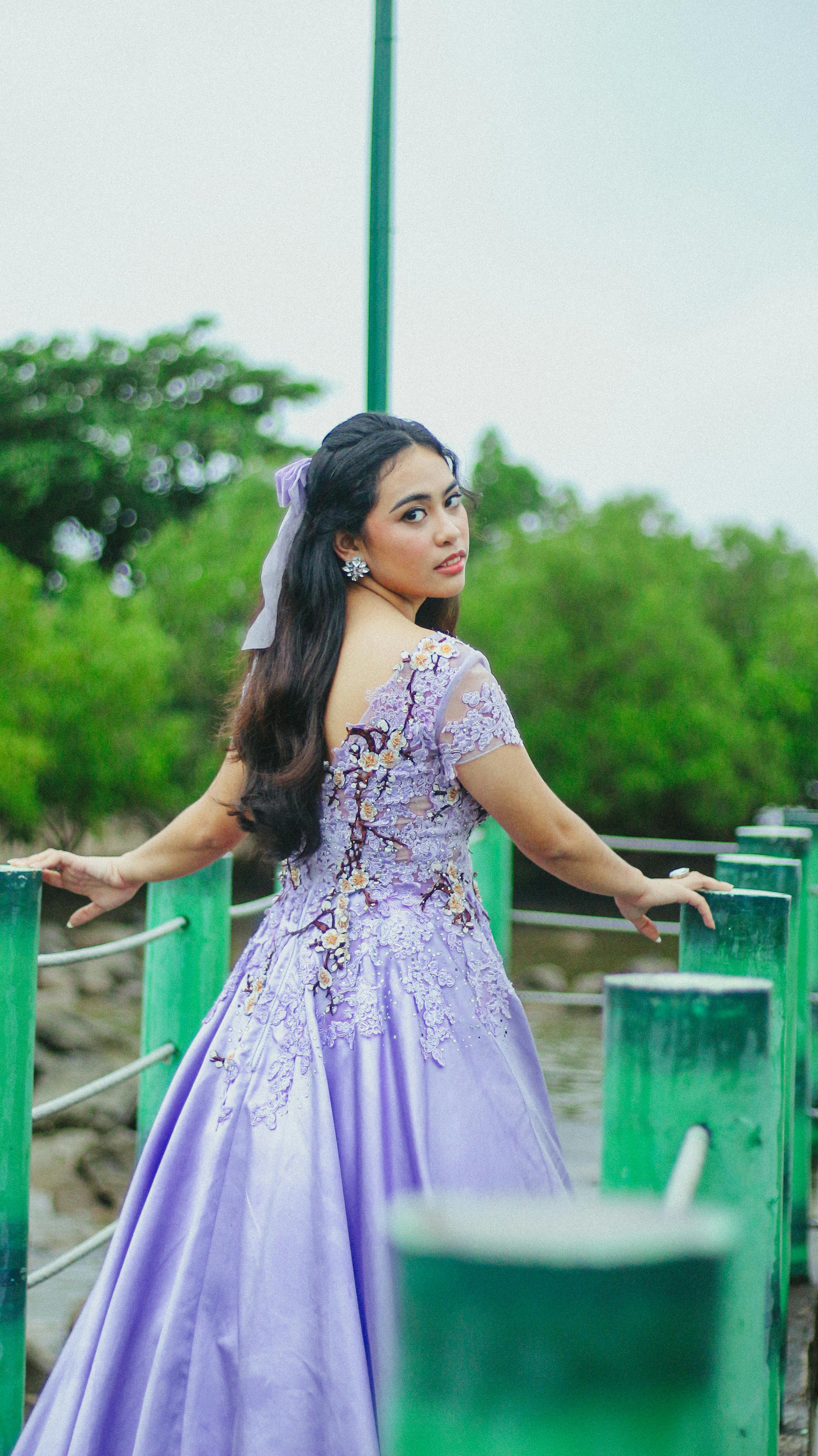 Young woman in lavender gown poses elegantly on an outdoor walkway with greenery.