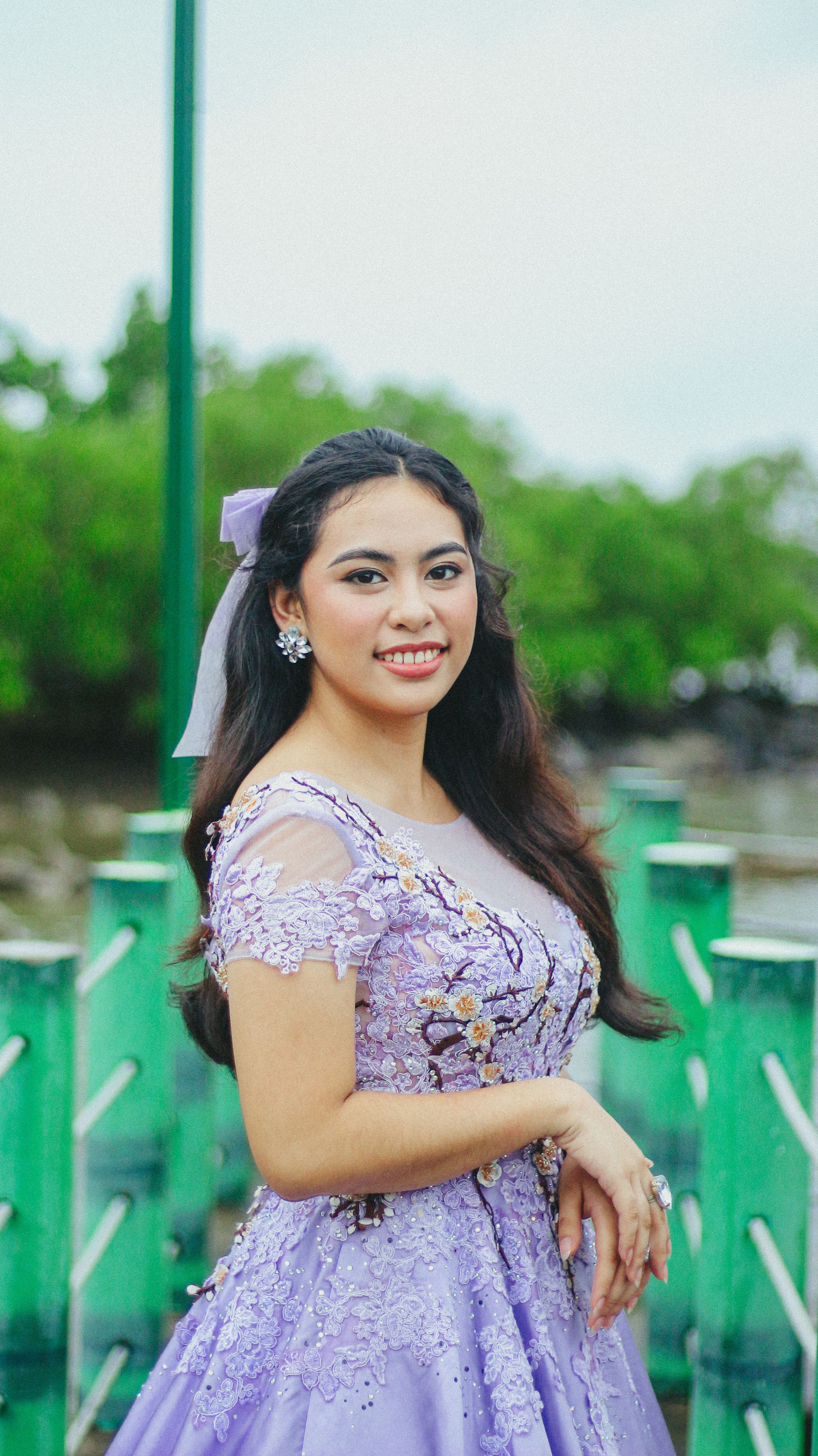 A young woman in a lavender gown poses gracefully outdoors with greenery background.
