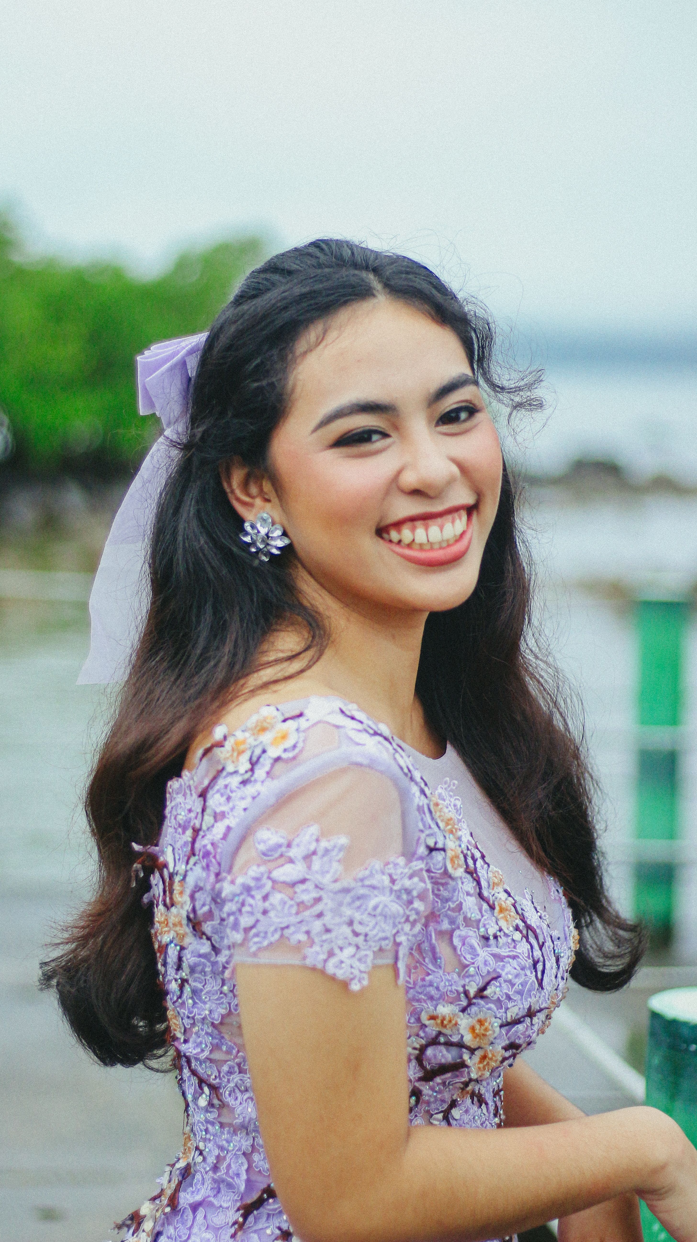 A young woman in a lavender floral dress smiles on a sunny day by the water.