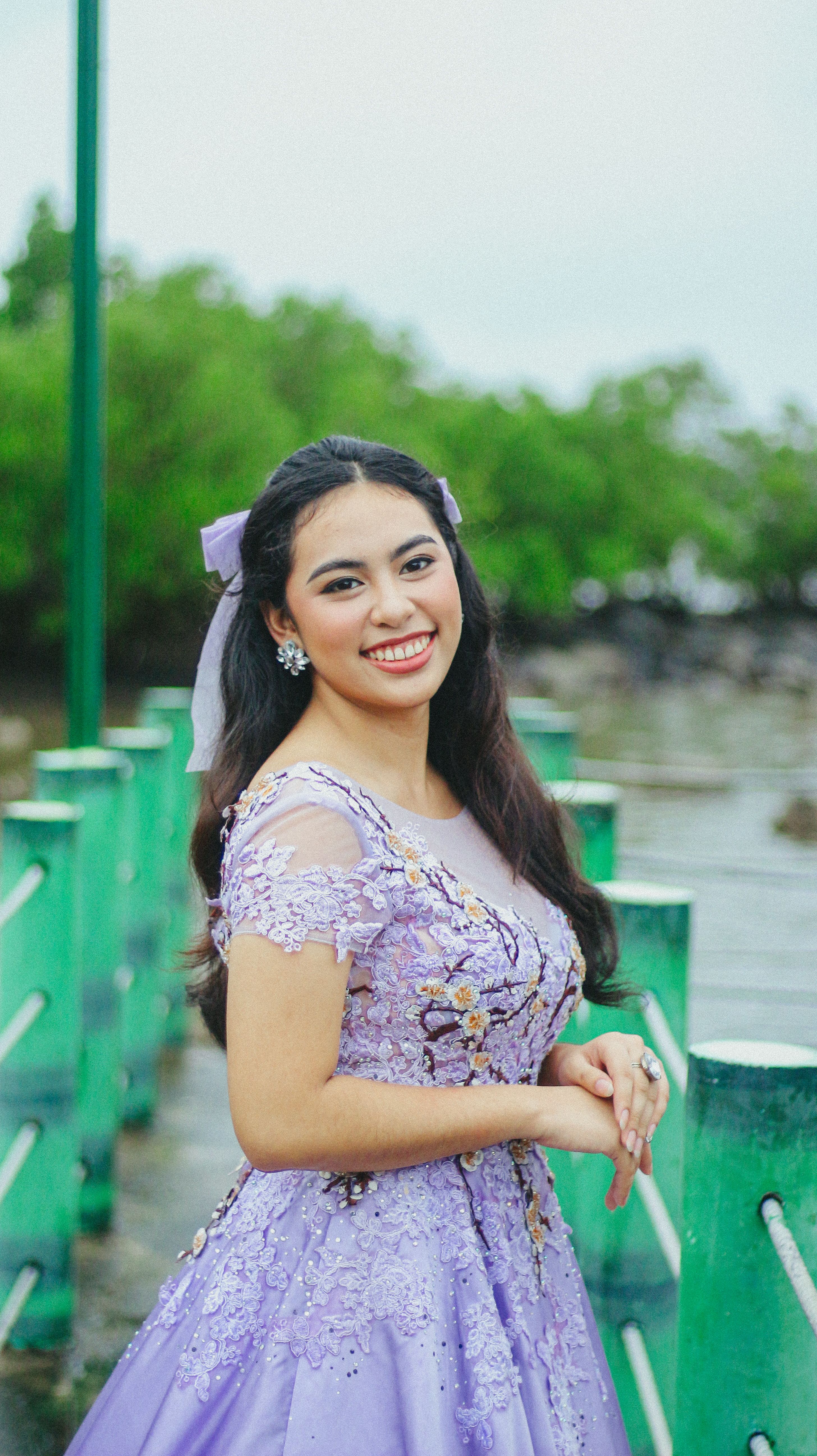 Smiling woman in a lavender dress posing outdoors on a bridge with greenery.
