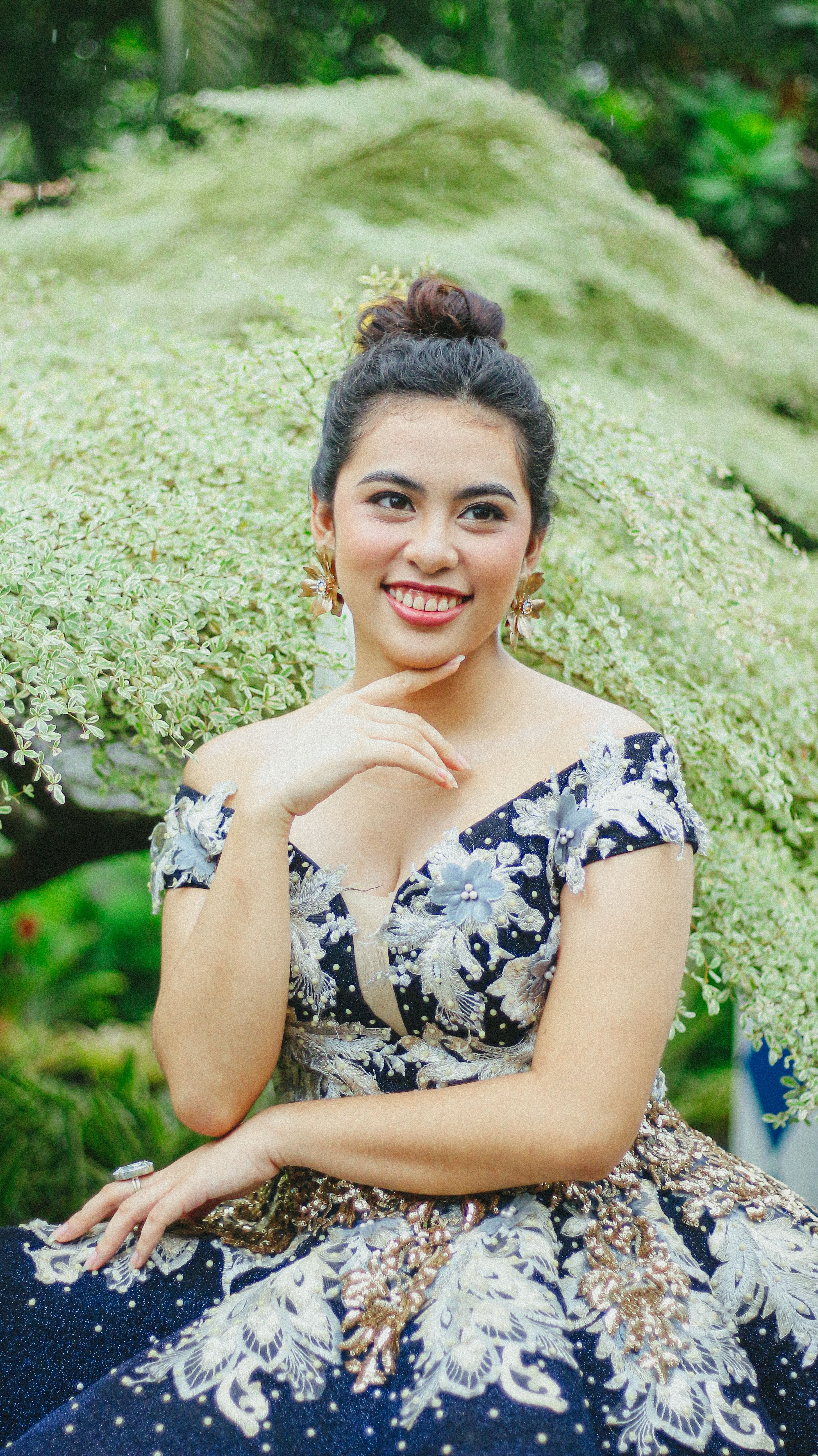 Smiling woman in ornate floral dress outdoors among greenery, exuding elegance and joy.